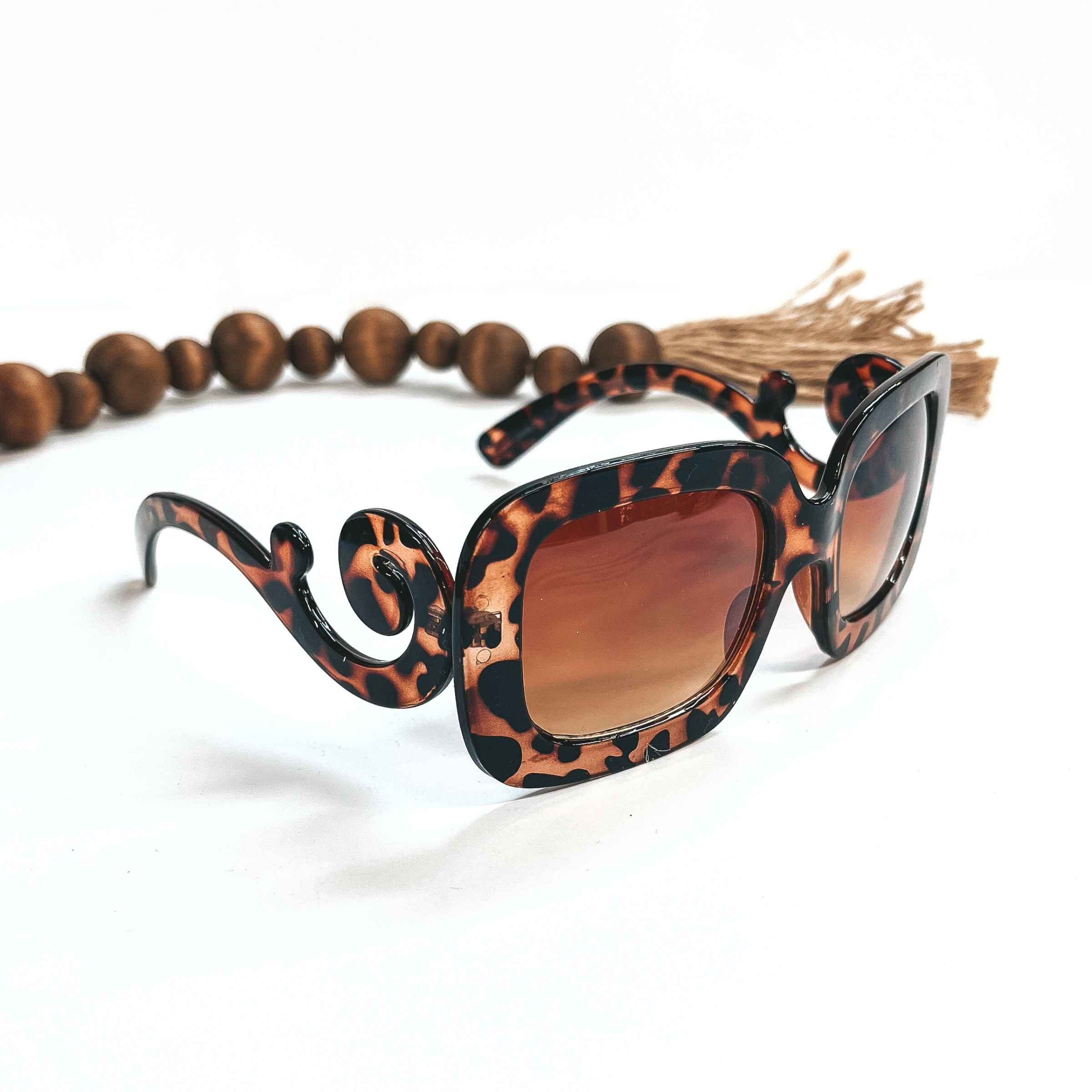 This a pair of tortouis print frame sunglasses, square rounded shape with a brown  lense. The sides of the sunglasses have a large swirls. These sunglasses are taken  on a white background with dark brown beads and brown tassel in the back as decor.