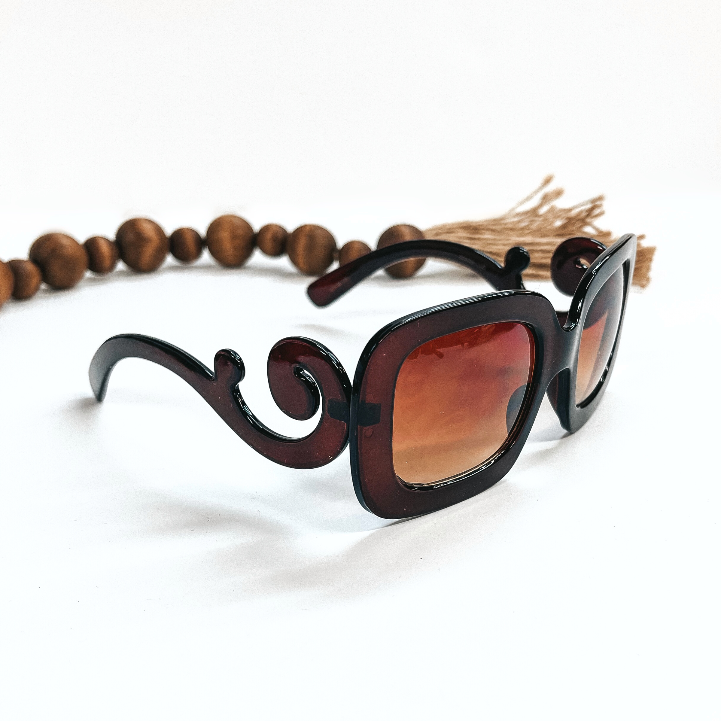 This a pair of brown frame sunglasses, square rounded shape with a brown  lense. The sides of the sunglasses have a large swirls. These sunglasses are taken  on a white background with dark brown beads and brown tassel in the back as decor.
