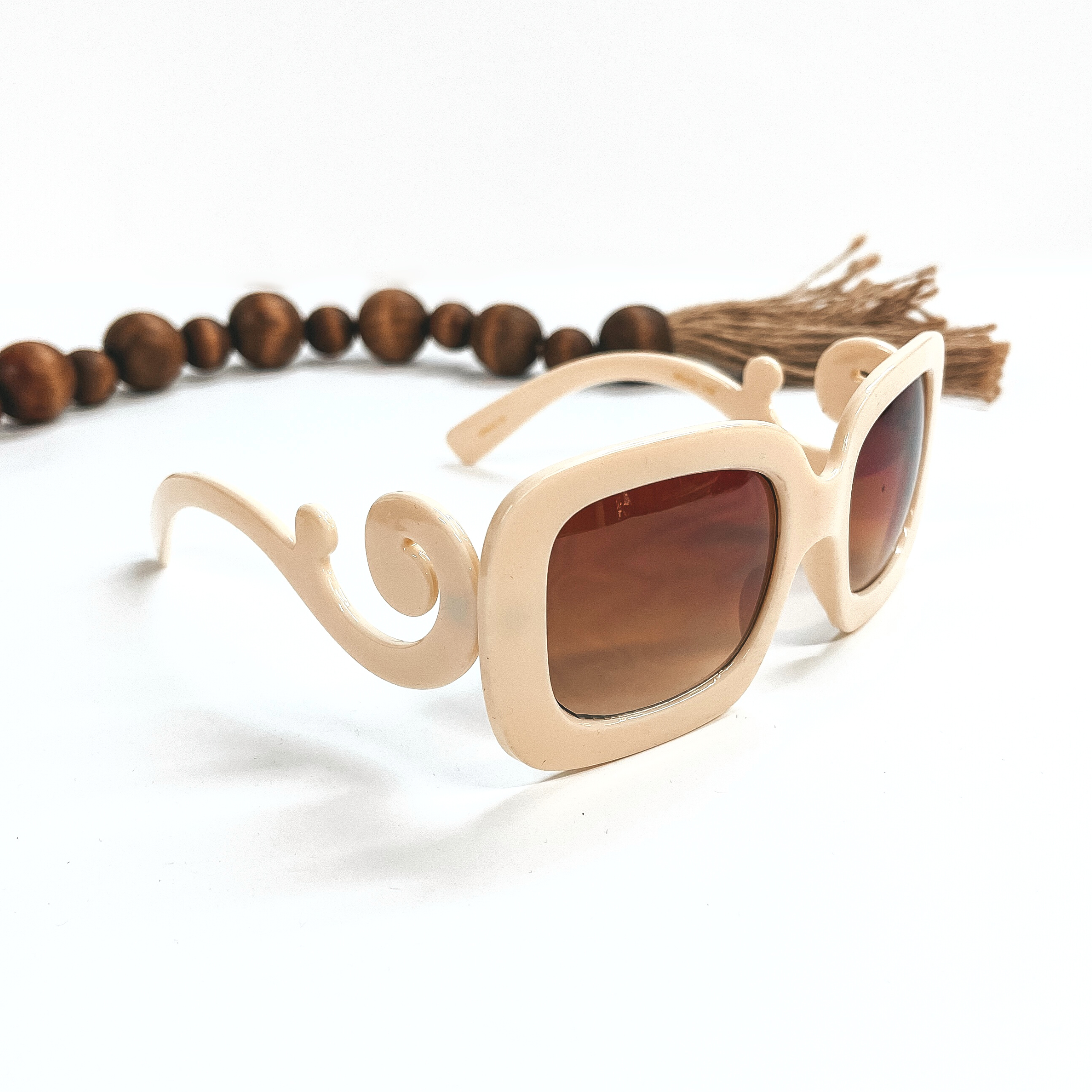 This a pair of cream frame sunglasses, square rounded shape with a brown  lense. The sides of the sunglasses have a large swirls. These sunglasses are taken  on a white background with dark brown beads and brown tassel in the back as decor.