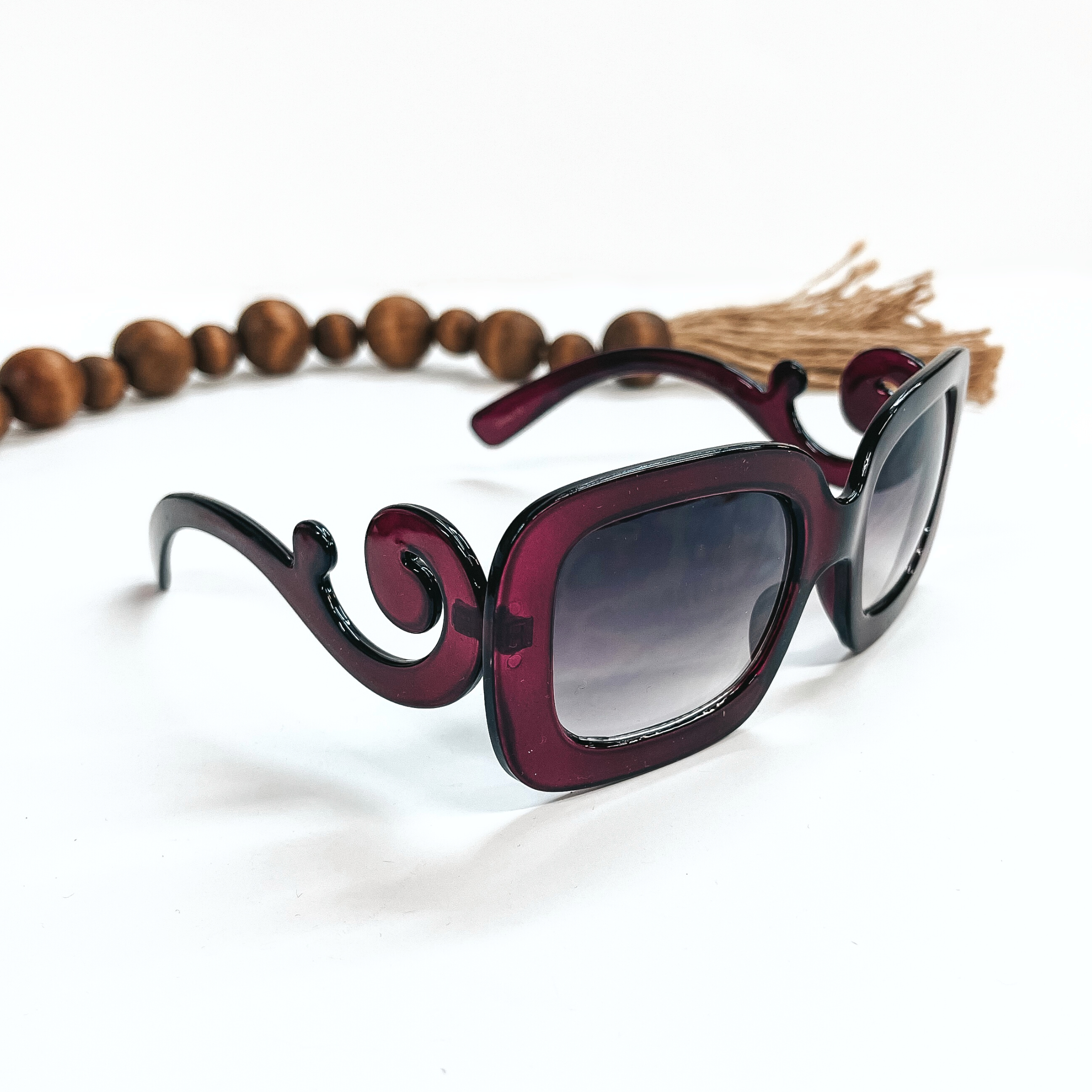 This is a pair of purple frame sunglasses, square rounded shape with a dark grey/black  lense. The sides of the sunglasses have a large swirls. These sunglasses are taken  on a white background with dark brown beads and brown tassel in the back as decor.