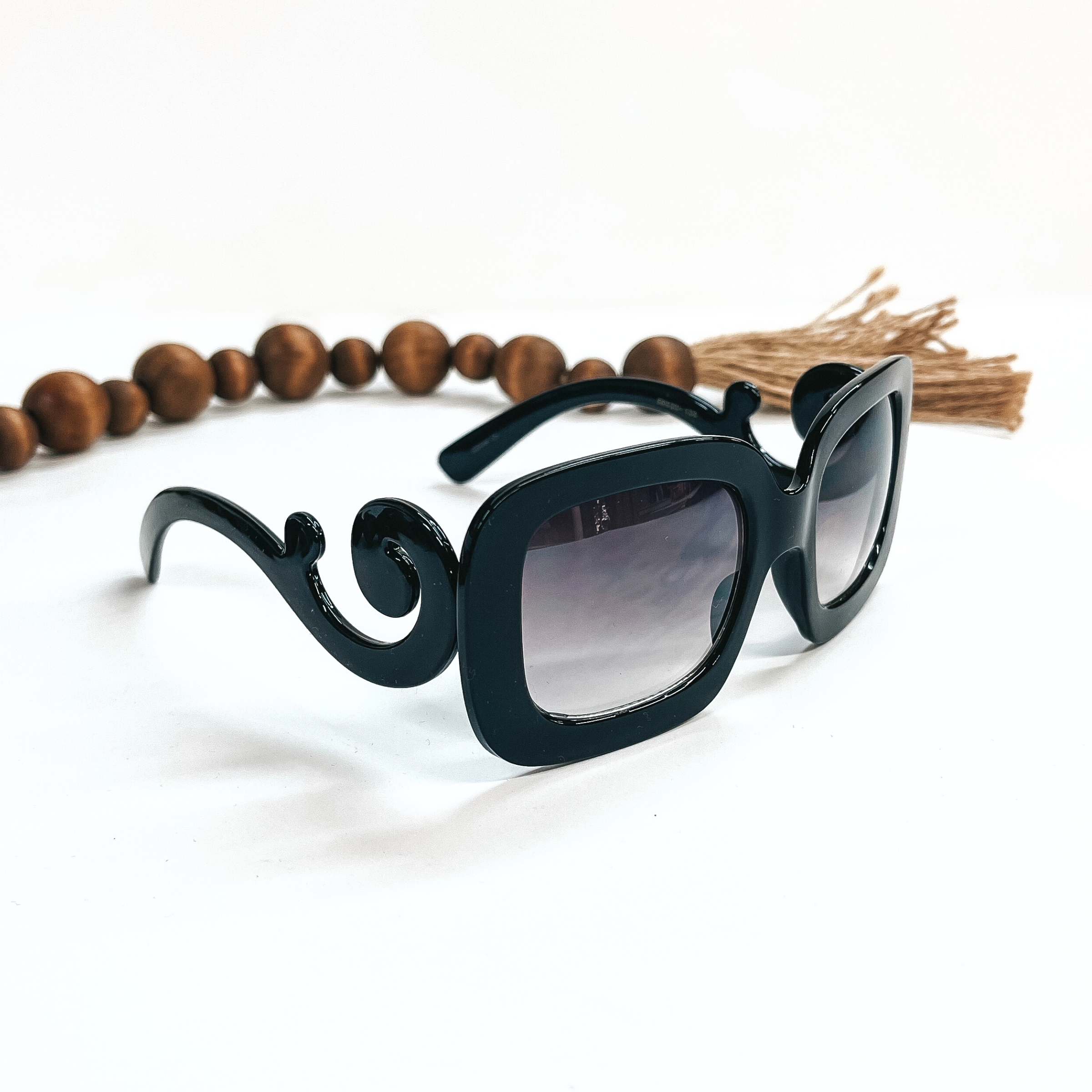 This is a pair of black frame sunglasses, square rounded shape with a dark grey/black  lense. The sides of the sunglasses have a large swirls. These sunglasses are taken  on a white background with dark brown beads and brown tassel in the back as decor.