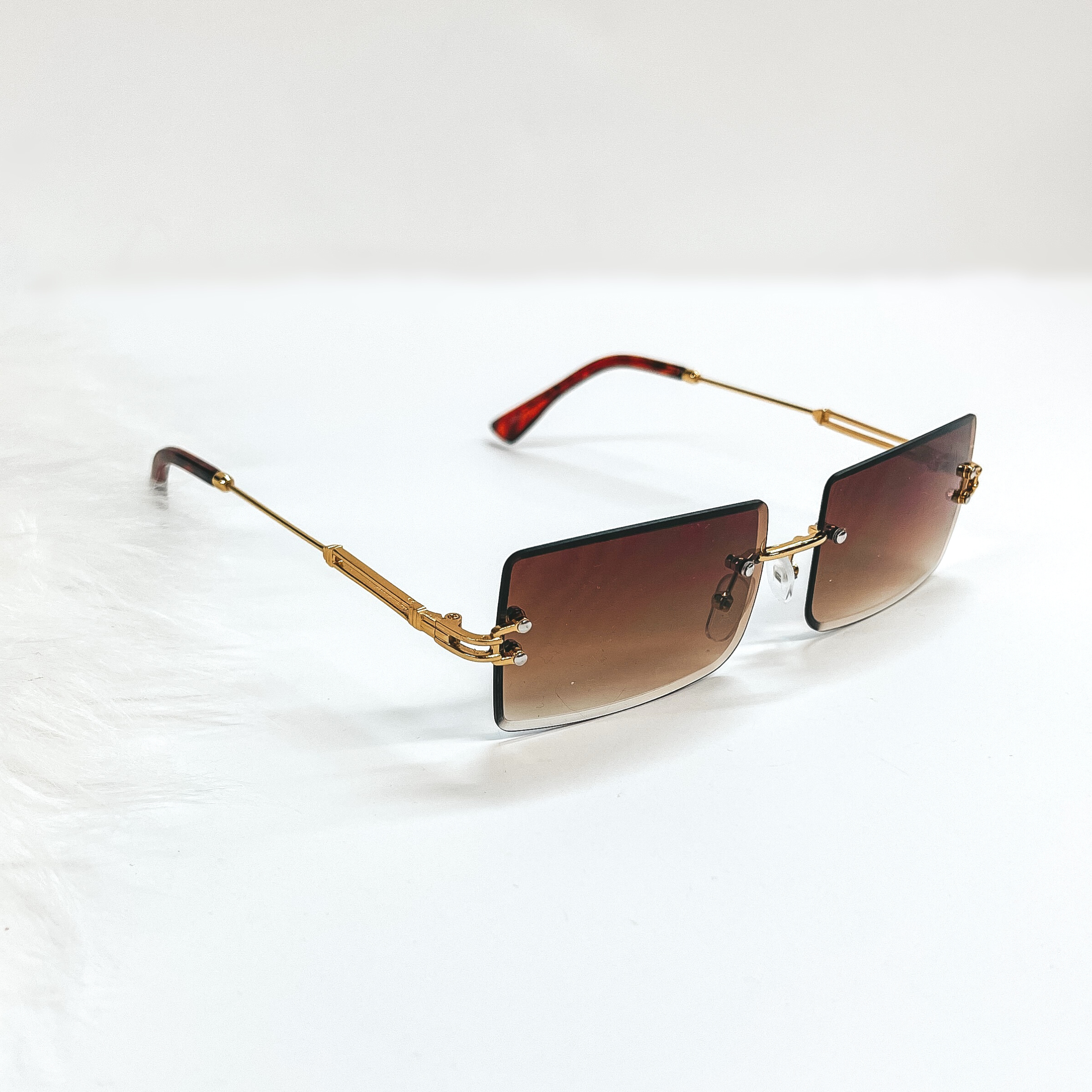 This is a brown pair of sunglasses, the lenses are brown with no frame and they have  gold connextors. The ear pieces are tortuoise print. These sunglasses are taken  on a white background with a piece of white fur in the side as decor.