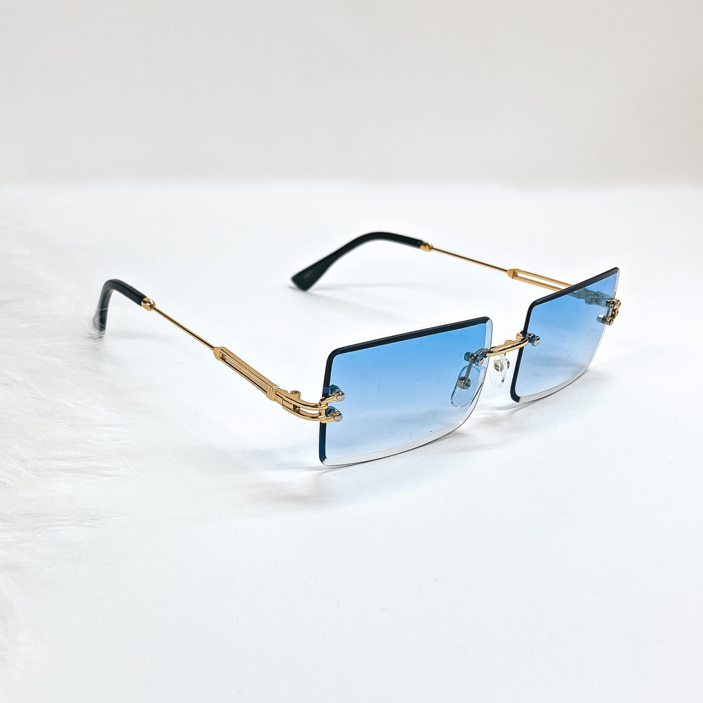 This is a blue pair of sunglasses, the lenses are blue with no frame and they have  gold connectors. The ear pieces are black. These sunglasses are taken  on a white background with a piece of white fur in the side as decor.