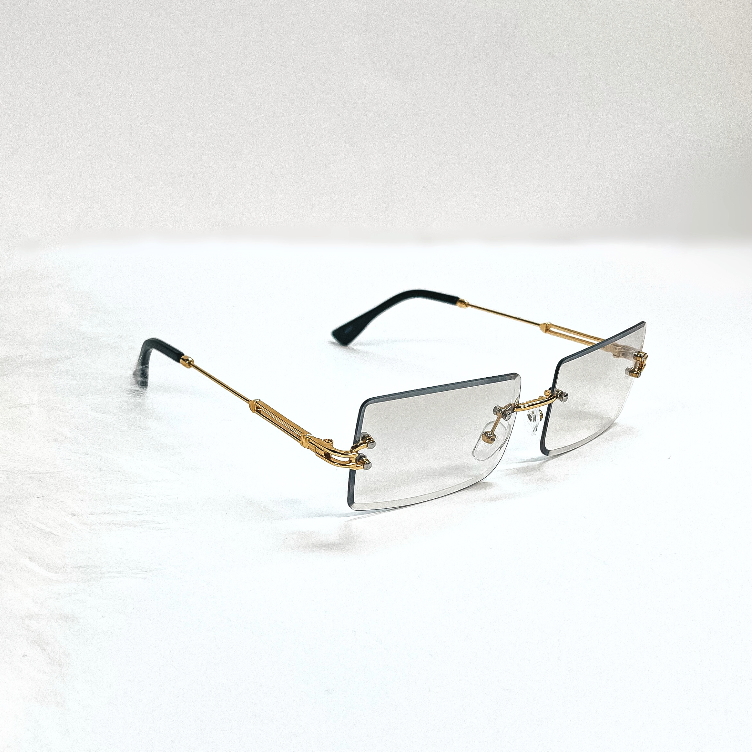 This is a clear grey/brown pair of sunglasses, the lenses are clear brown/grey  with no frame and they have  gold connectors. The ear pieces are black. These sunglasses are taken  on a white background with a piece of white fur in the side as decor.