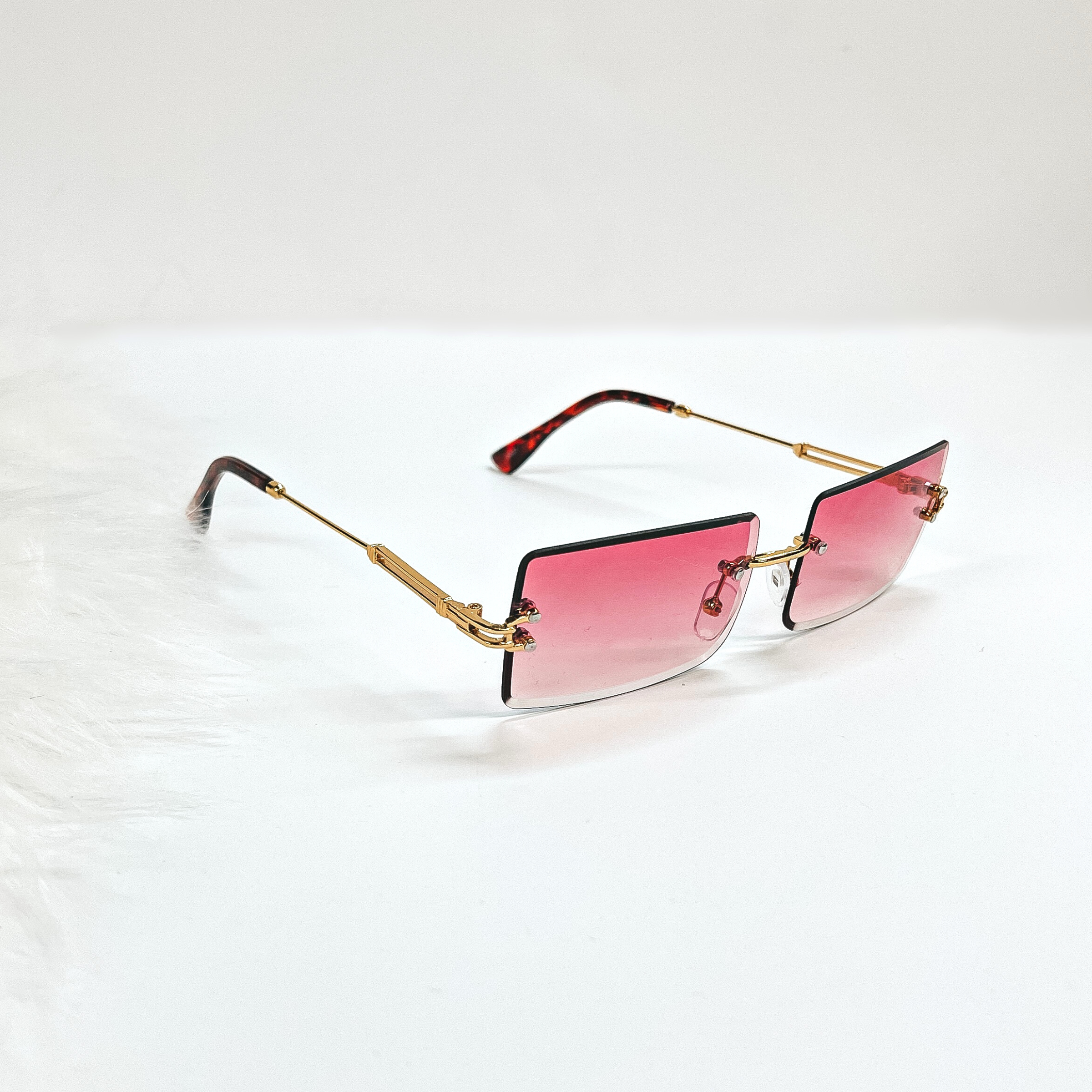 This is a pink pair of sunglasses, the lenses are pink  with no frame and they have  gold connectors. The ear pieces are tortuoise print. These sunglasses are taken  on a white background with a piece of white fur in the side as decor.