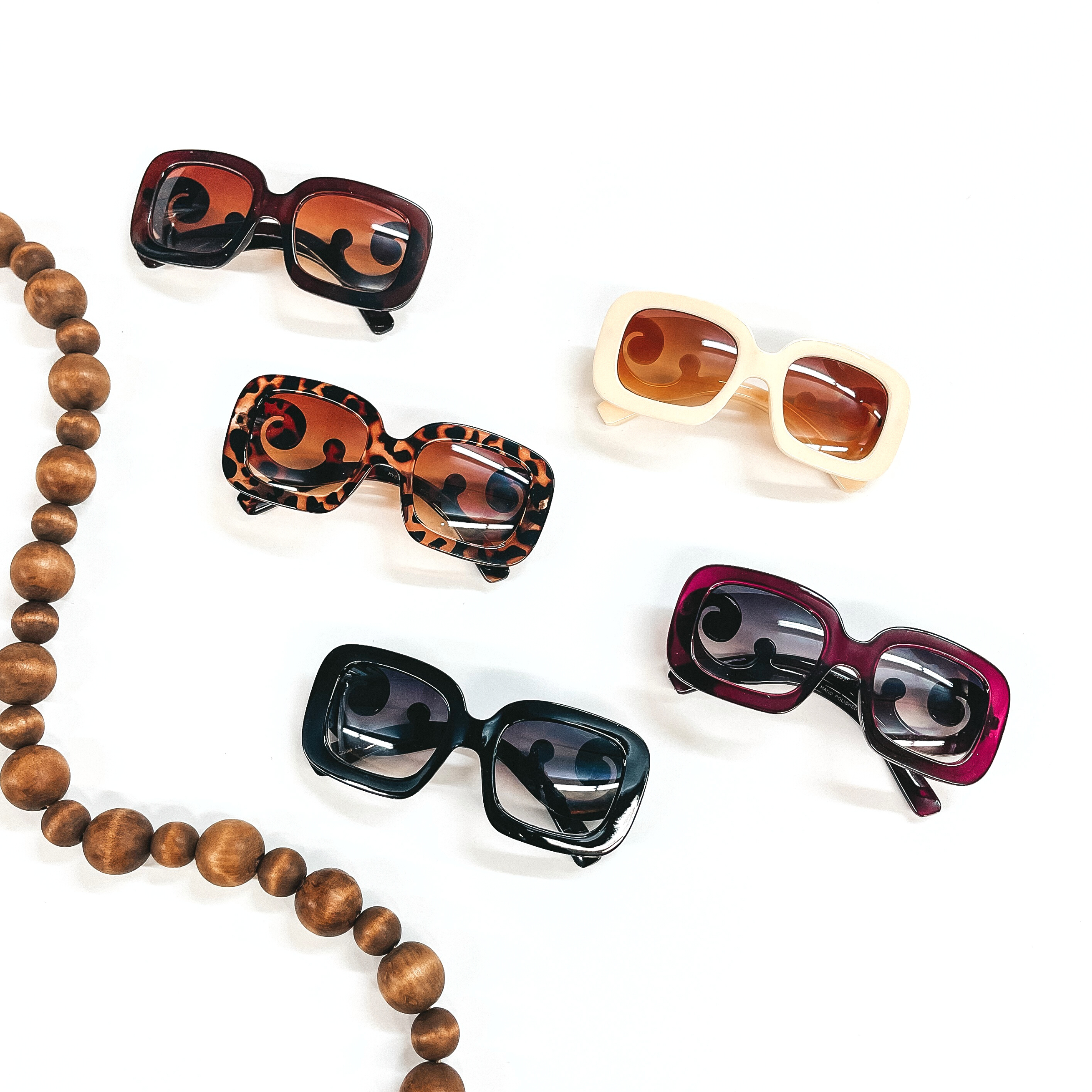 These are five pairs of large square rounded sunglasses in various colors and prints. There is brown, cream, tortuoise print, purple, and black. These sunglasses are  taken on a white background with brown beads in the side as decor.