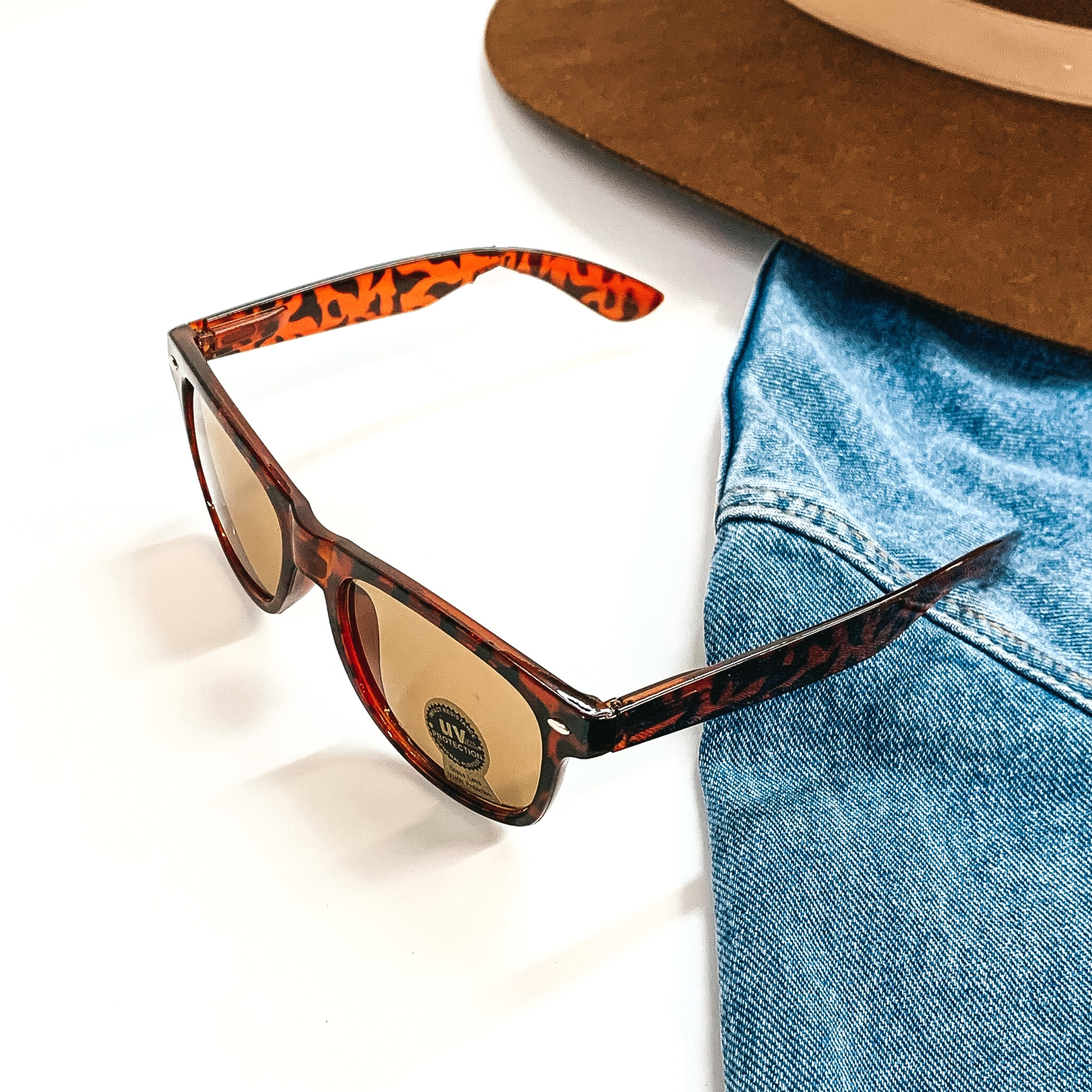This pair of sunglasses has a brown tortouise print frame with brown  lenses and silver detailing  in the side. This pair of sunglasses are taken on a white background and jean jacket  sleeve with a brown felt hat as decor.