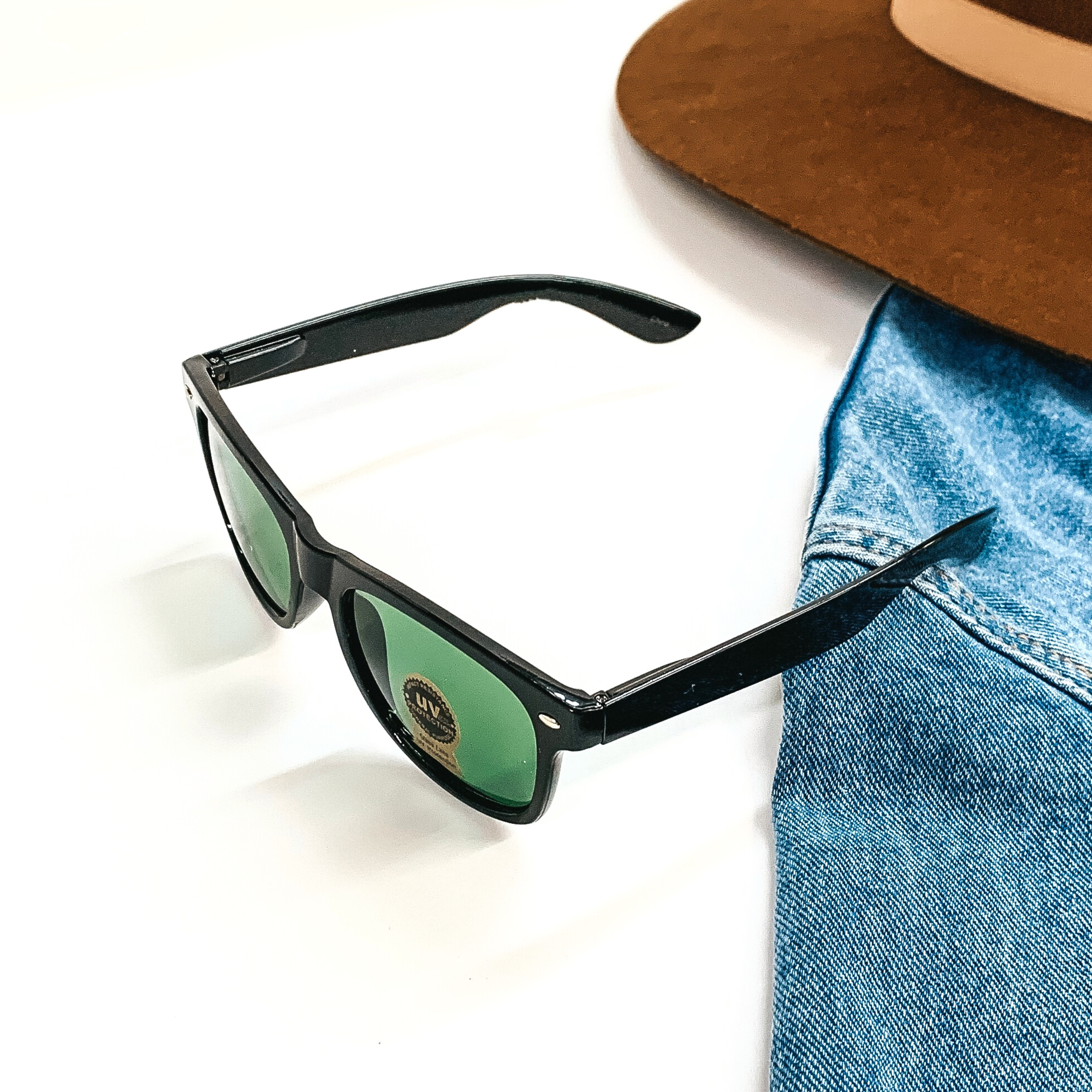 This pair of sunglasses has a black frame with dark green lenses and silver detailing  in the side. This pair of sunglasses are taken on a white background and jean jacket  sleeve with a brown felt hat as decor.