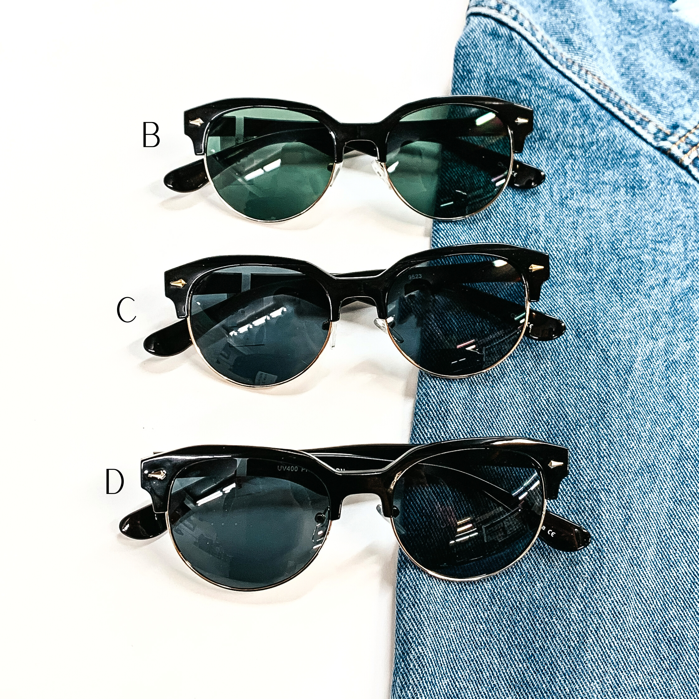 There are three pairs of browline style sunglasses in black for the frame  and different colors of lenses. Style B has a black frame, dark green  lense with a gold tone outline. Style C has a black frame, black/dark grey lense with a  gold tone outline. Style D has a black frame with a black/dark grey lense  with a silver tone outline. These three pairs of sunglasses are taken on a white  background and on a jean jacket sleeve.