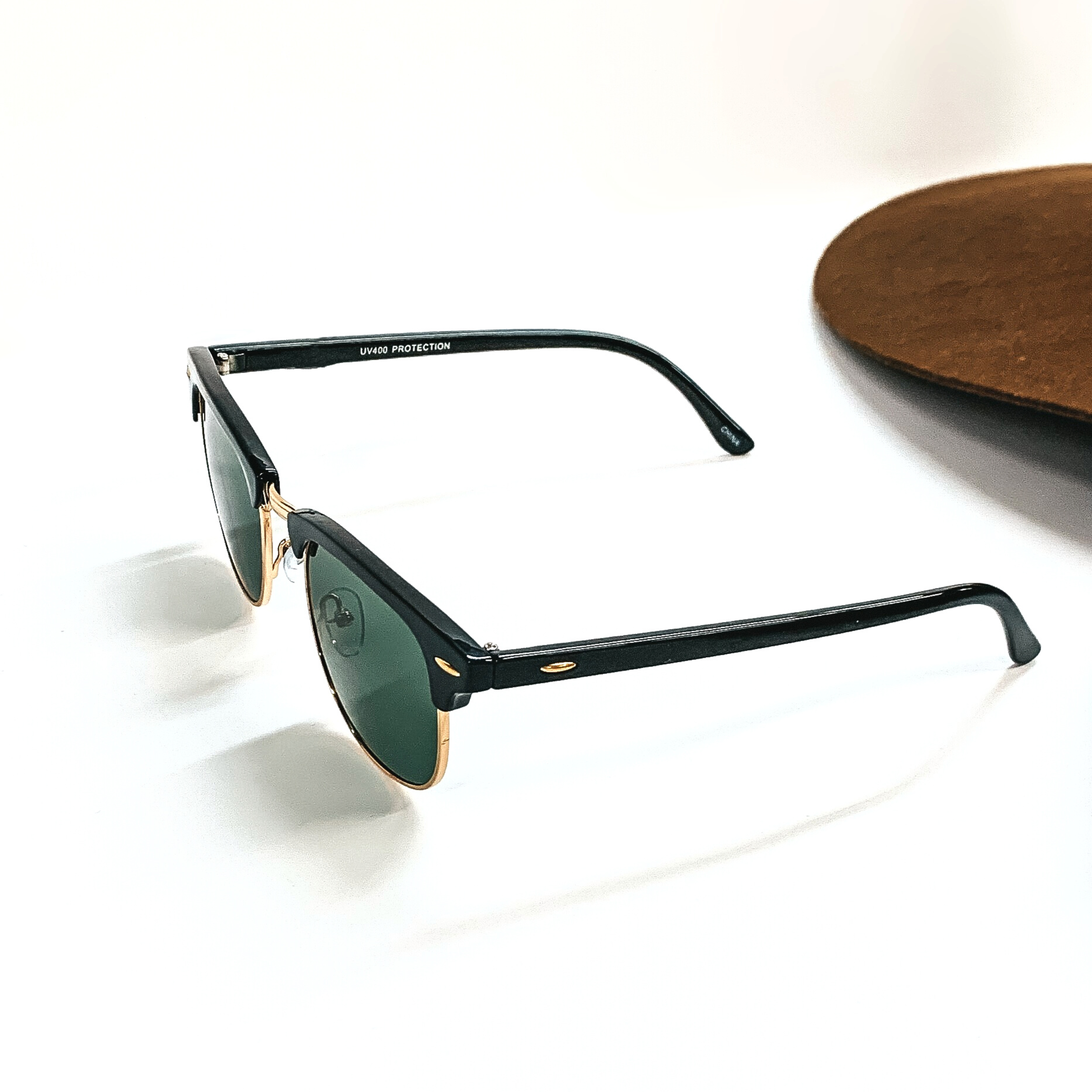 This pair of browline sunglasses have a black frame, dark green lense, with a gold tone  outline. These sunglasses are taken on a white background with a dark brown felt hat in  the back as decor.