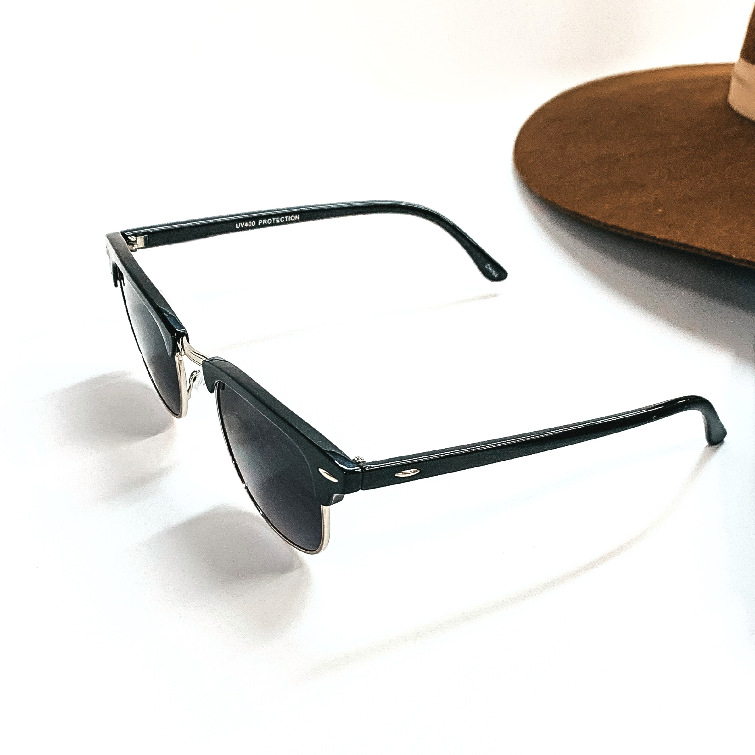 This pair of browline sunglasses have a black frame, black/dark grey lense, with a silver tone  outline. These sunglasses are taken on a white background with a dark brown felt hat in  the back as decor.