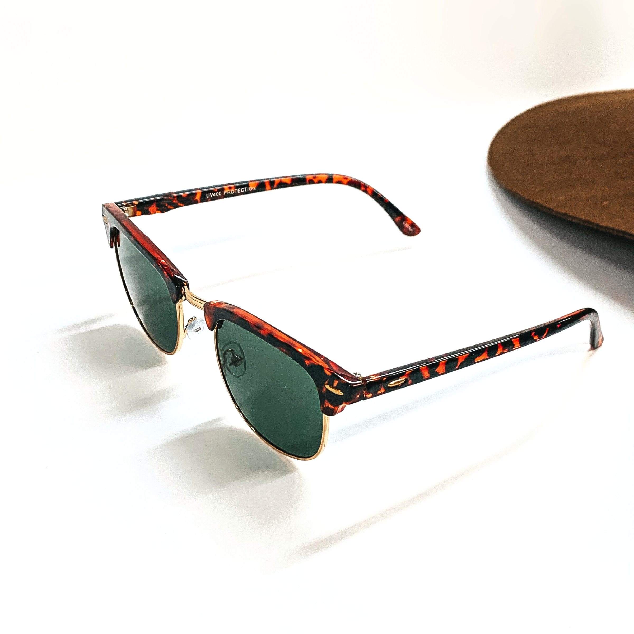 This pair of browline sunglasses have a tortuoise print frame, dark green lense, with a gold tone  outline. These sunglasses are taken on a white background with a dark brown felt hat in  the back as decor.
