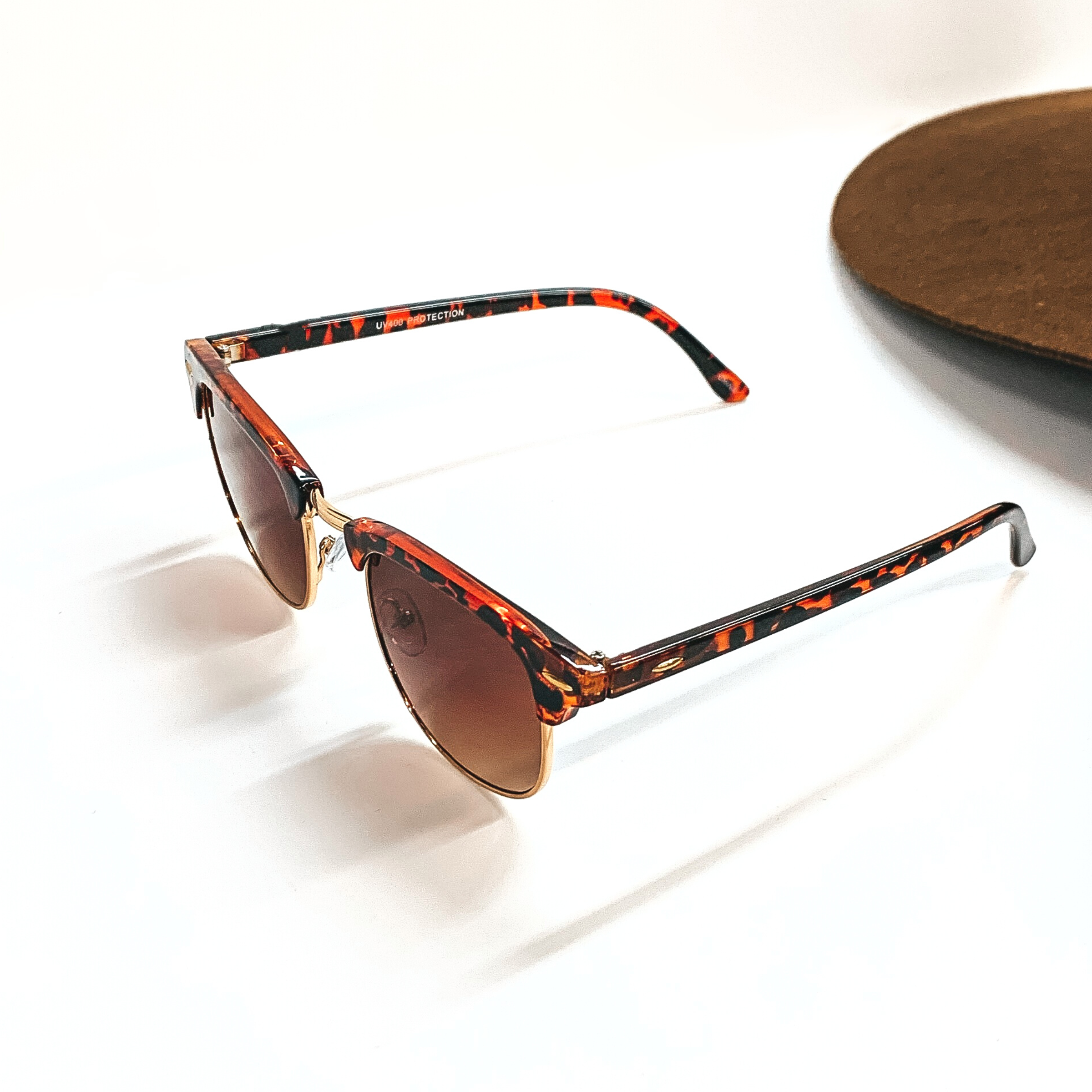 This pair of browline sunglasses have a tortuoise print frame, brown lense, with a gold tone  outline. These sunglasses are taken on a white background with a dark brown felt hat in  the back as decor.