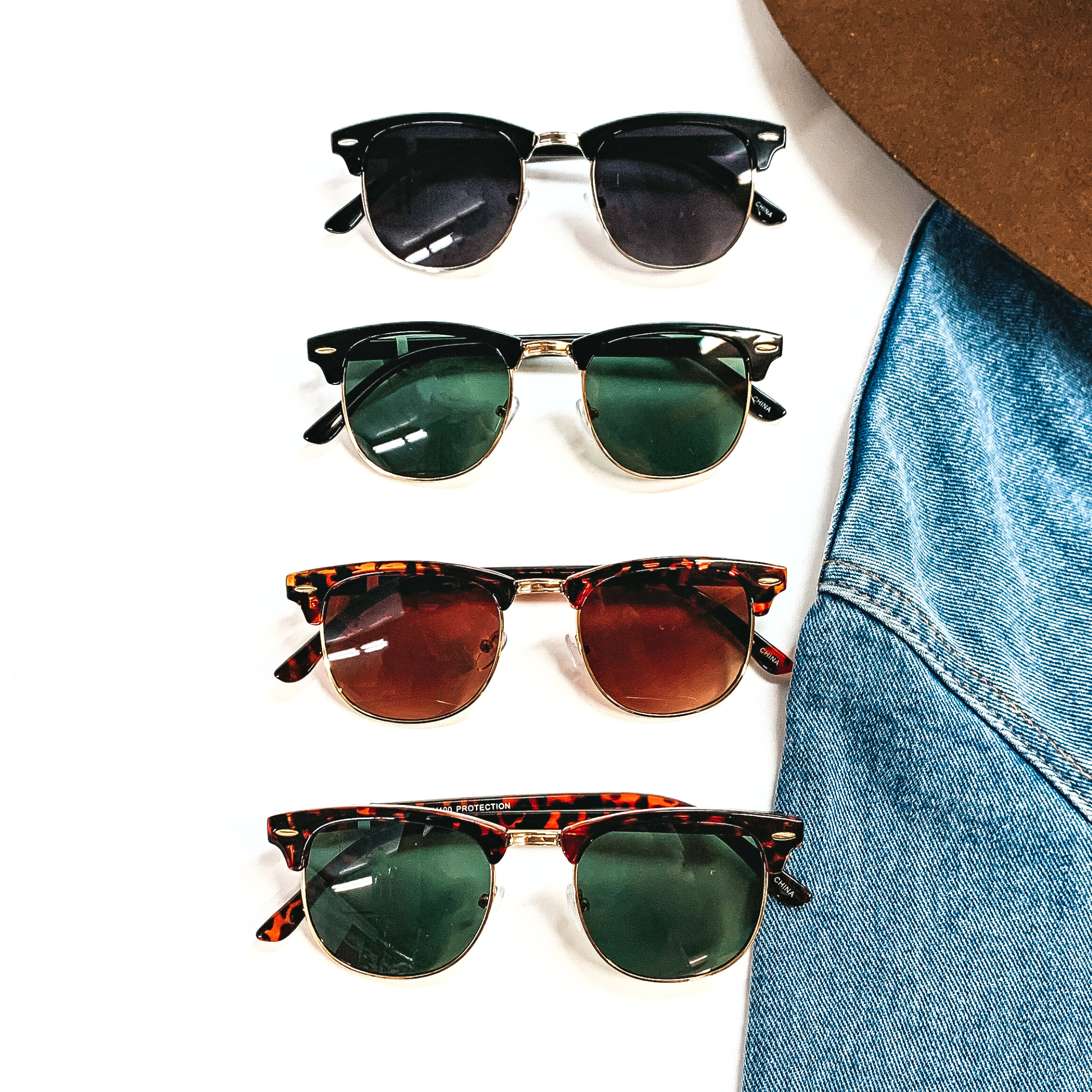 There are four pairs of sunglasses in different frame colors/print and color lenses. These sunglasses are taken on a white background and a jean jacket sleeve with a  dark brown felt hat on the top as decor.