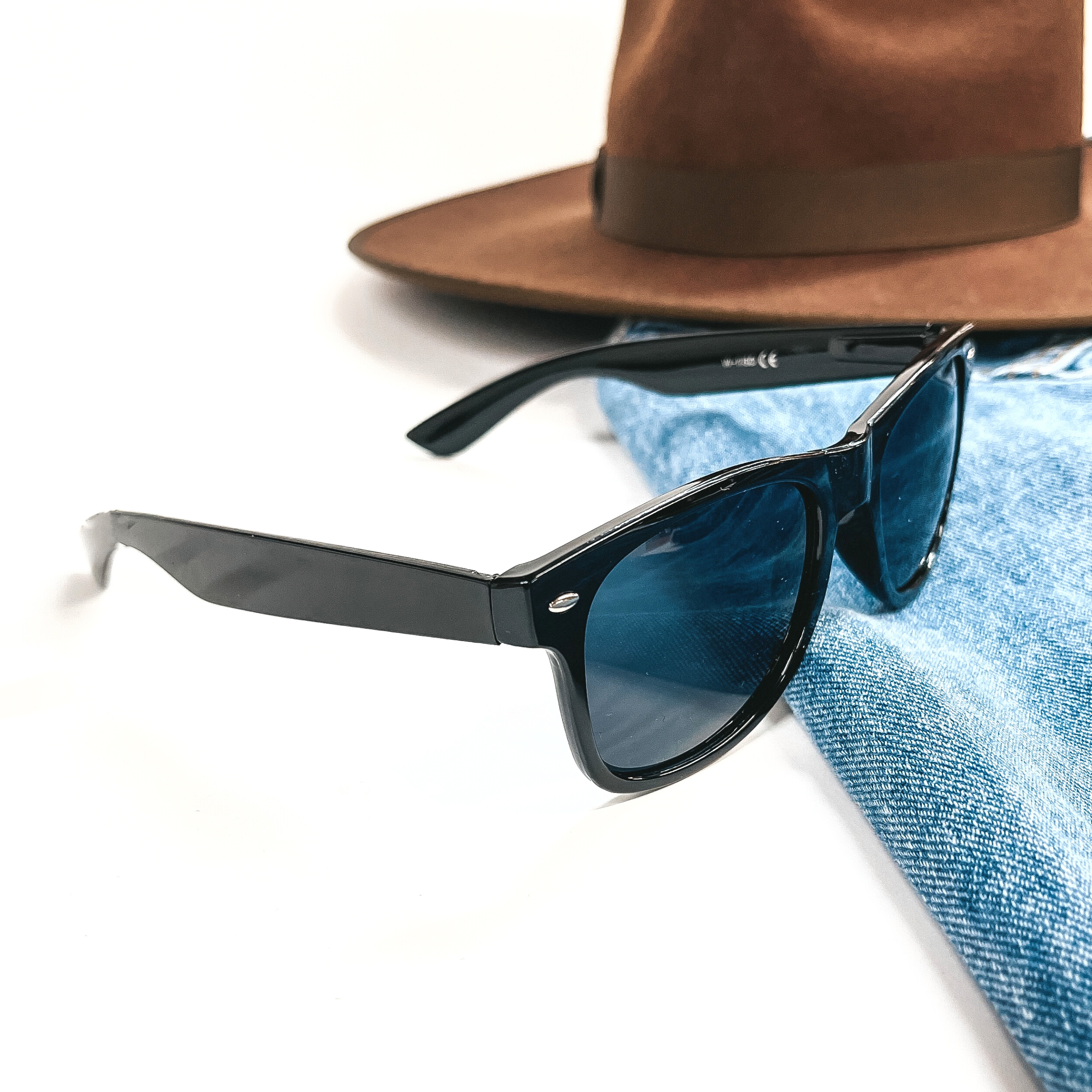 This is a black pair of sunglasses, the lenses are black/dark grey with silver  detailing in the side. These sunglasses are taken on a jean jacket sleeve with a  dark brown felt hat in the top as decor.