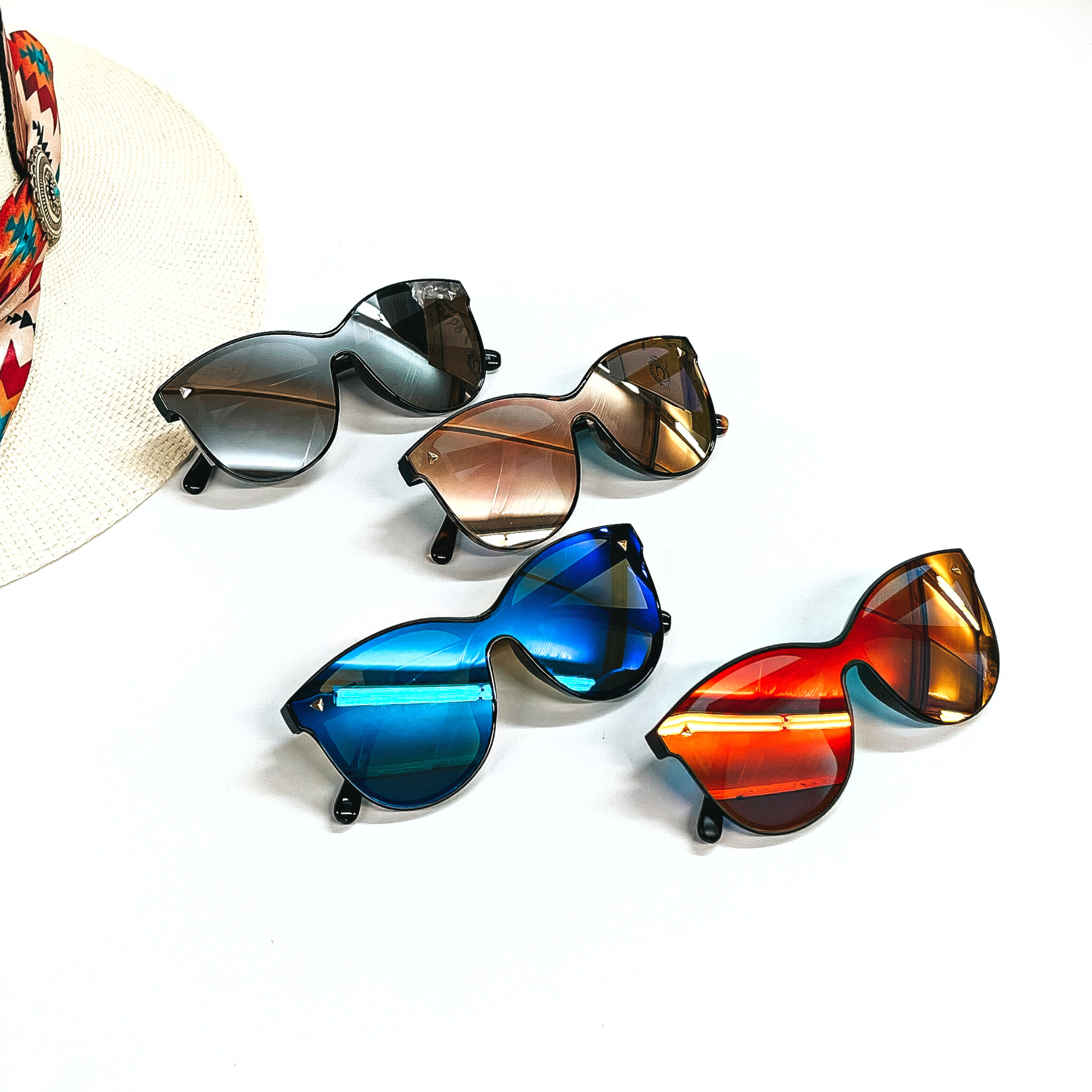 There are four pairs of medium shielf sunglasses in different colors such as a orange,  blue, coral, and silver. These sunglasses are taken on a white background with an ivory  straw hat and colorful aztec orint hat band as decor.