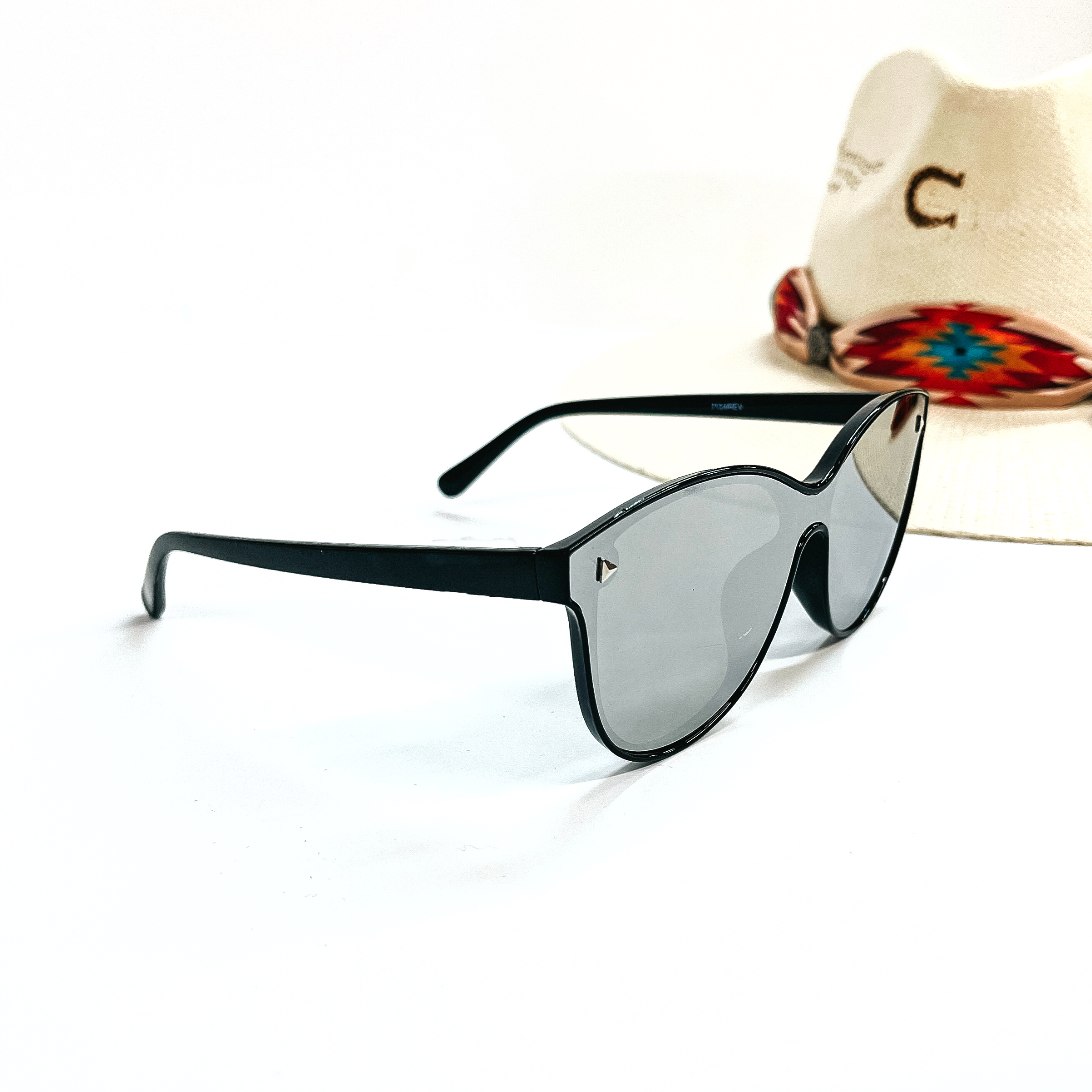 This is a pair of black matte frame sunglasses with a silver  mirrored lense and silver tone  detailing. This pair of sunglasses are taken on a white background and an ivory straw hat  with a colorful aztec print hat band in the back as  decor.