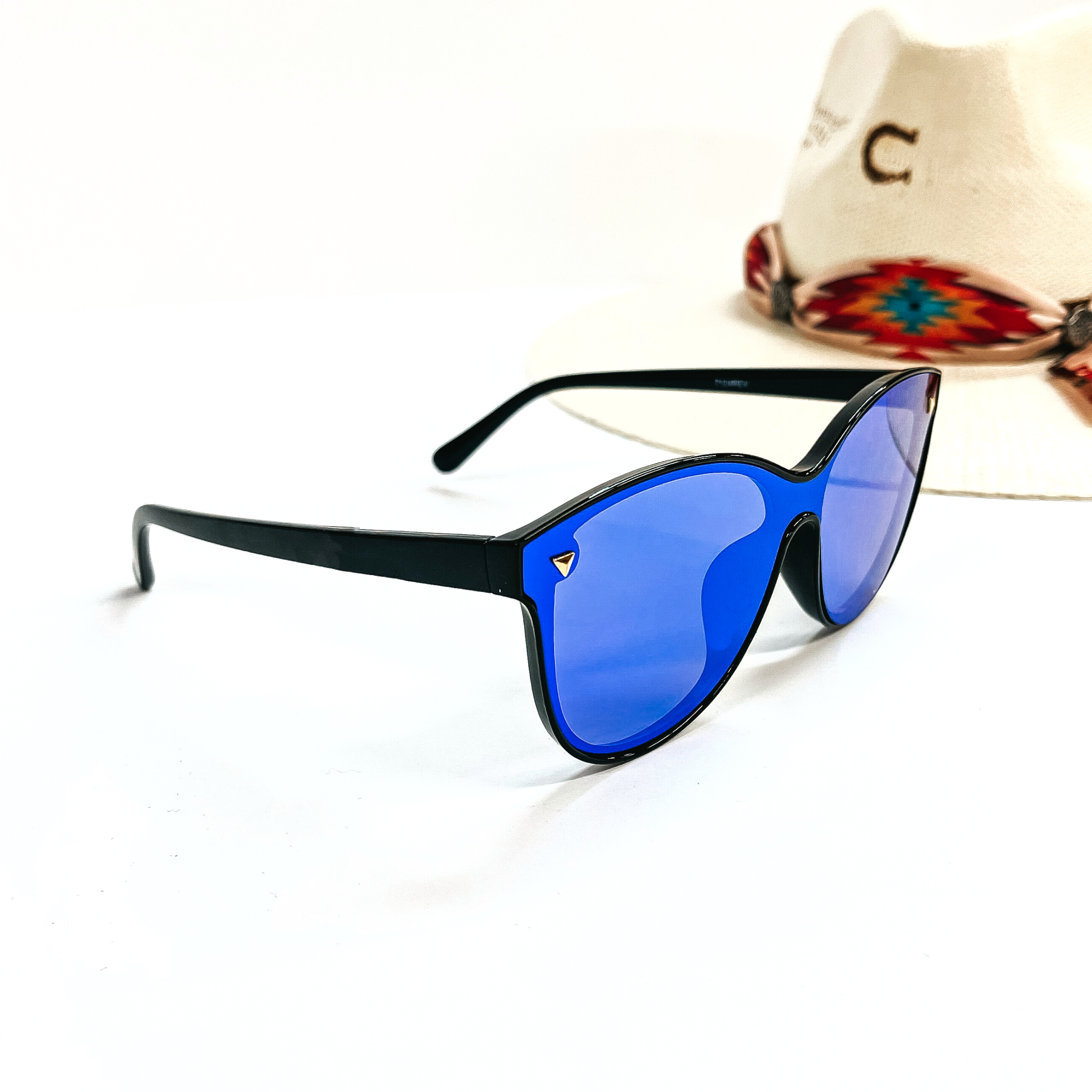 This is a pair of black frame sunglasses with a blue mirrored lense and silver tone  detailing. This pair of sunglasses are taken on a white background and an ivory straw hat  with a colorful aztec print hat band in the back as  decor.