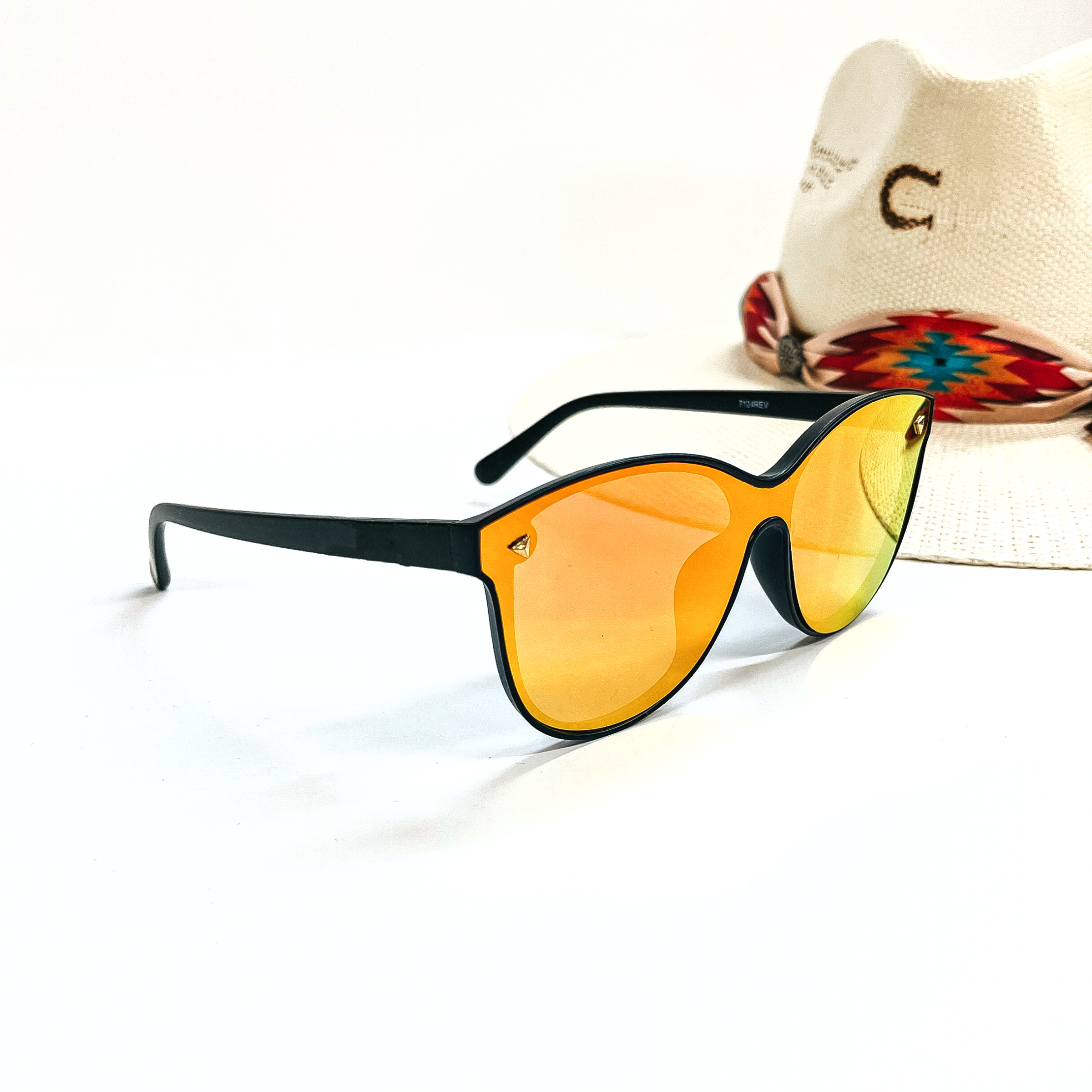 This is a pair of black matte frame sunglasses with an orange/red  mirrored lense and silver tone  detailing. This pair of sunglasses are taken on a white background and an ivory straw hat  with a colorful aztec print hat band in the back as  decor.
