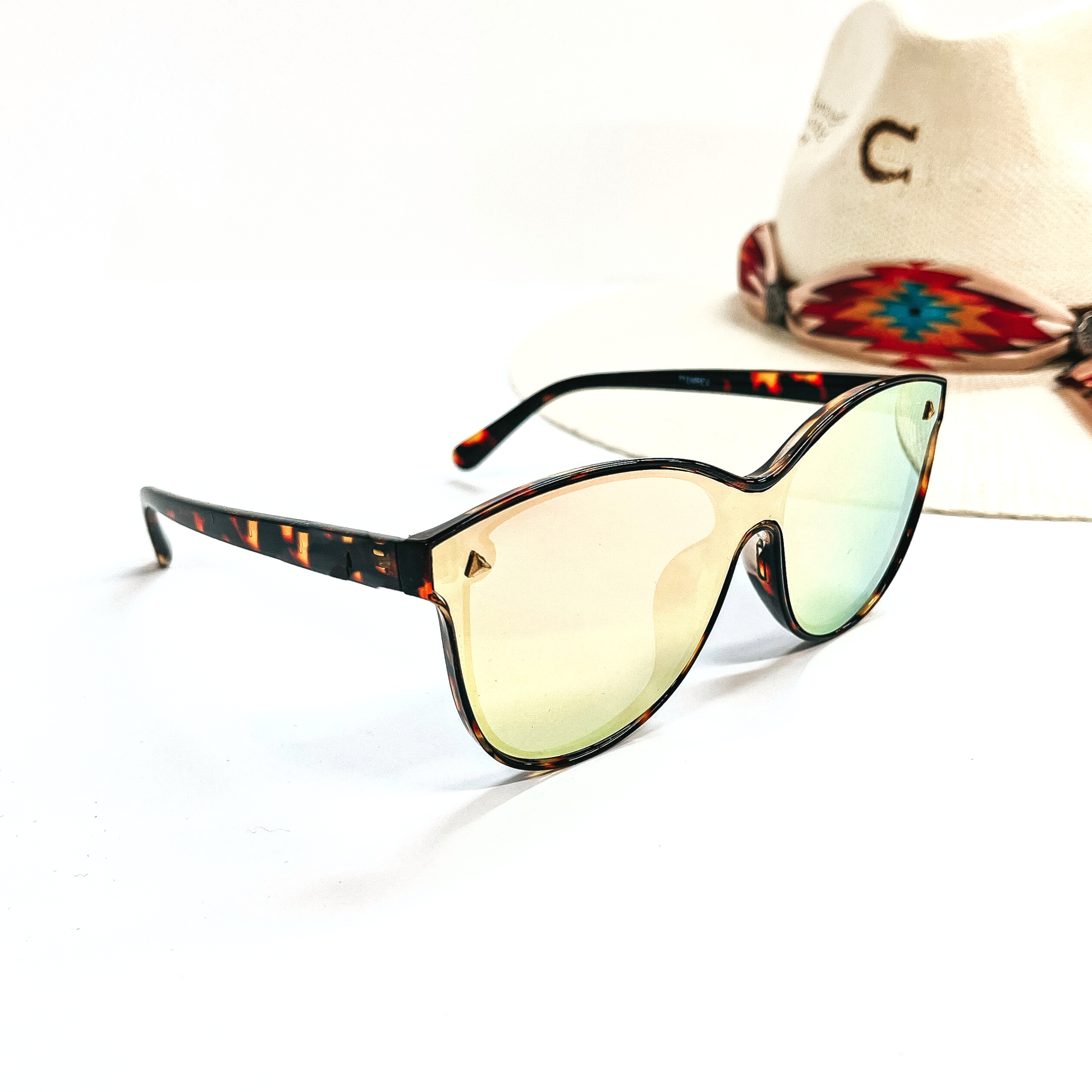 This is a pair of tortouise print  frame sunglasses with a green/coral/yellow  mirrored lense and silver tone  detailing. This pair of sunglasses are taken on a white background and an ivory straw hat  with a colorful aztec print hat band in the back as  decor.