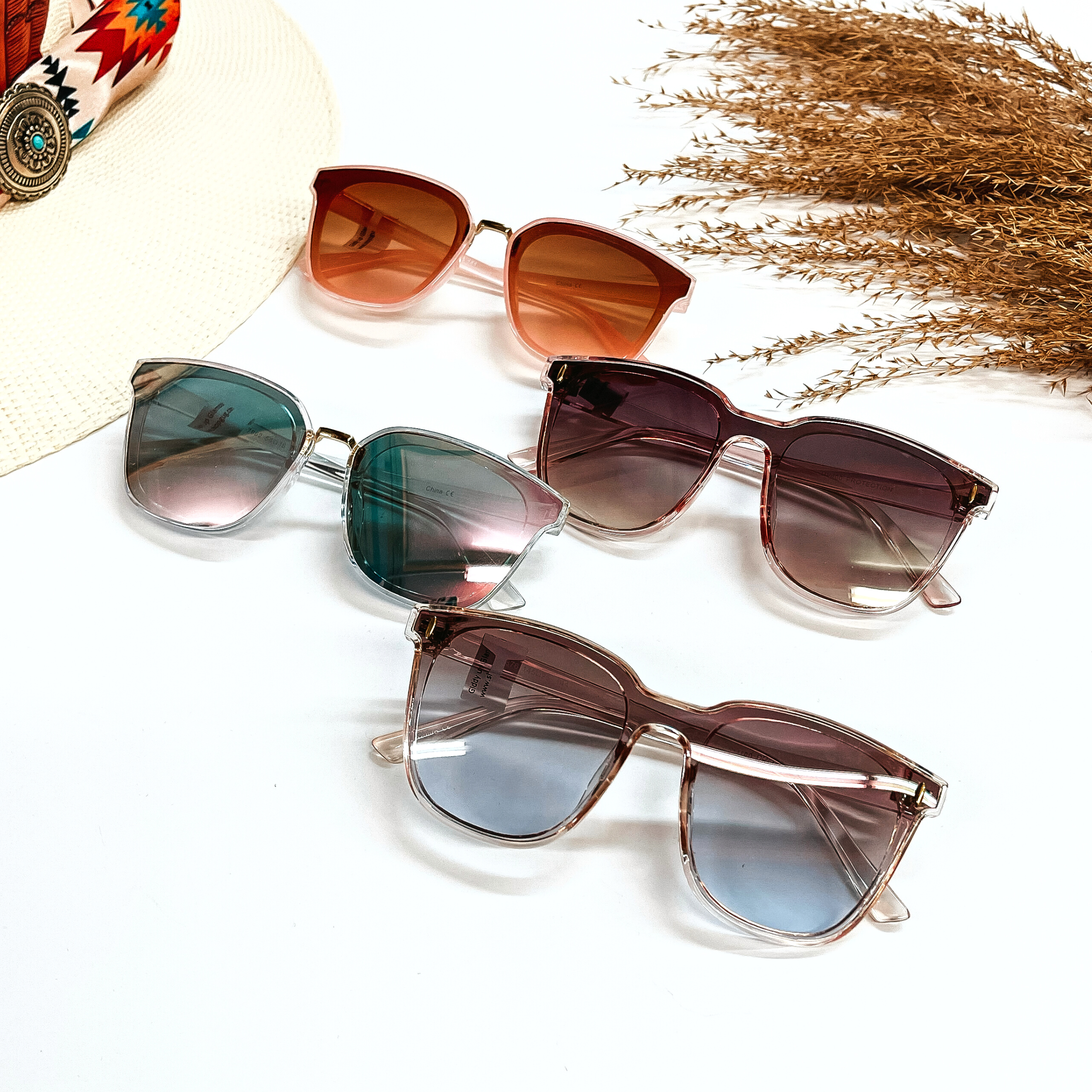 There are four pairs of wayfarer style sunglasses in different shades of pink/rose gold  frames and lenses. These pair of sunglasses are taken on a white background with a  straw hat and a colorful aztec print in the side, and a brown plant as decor.
