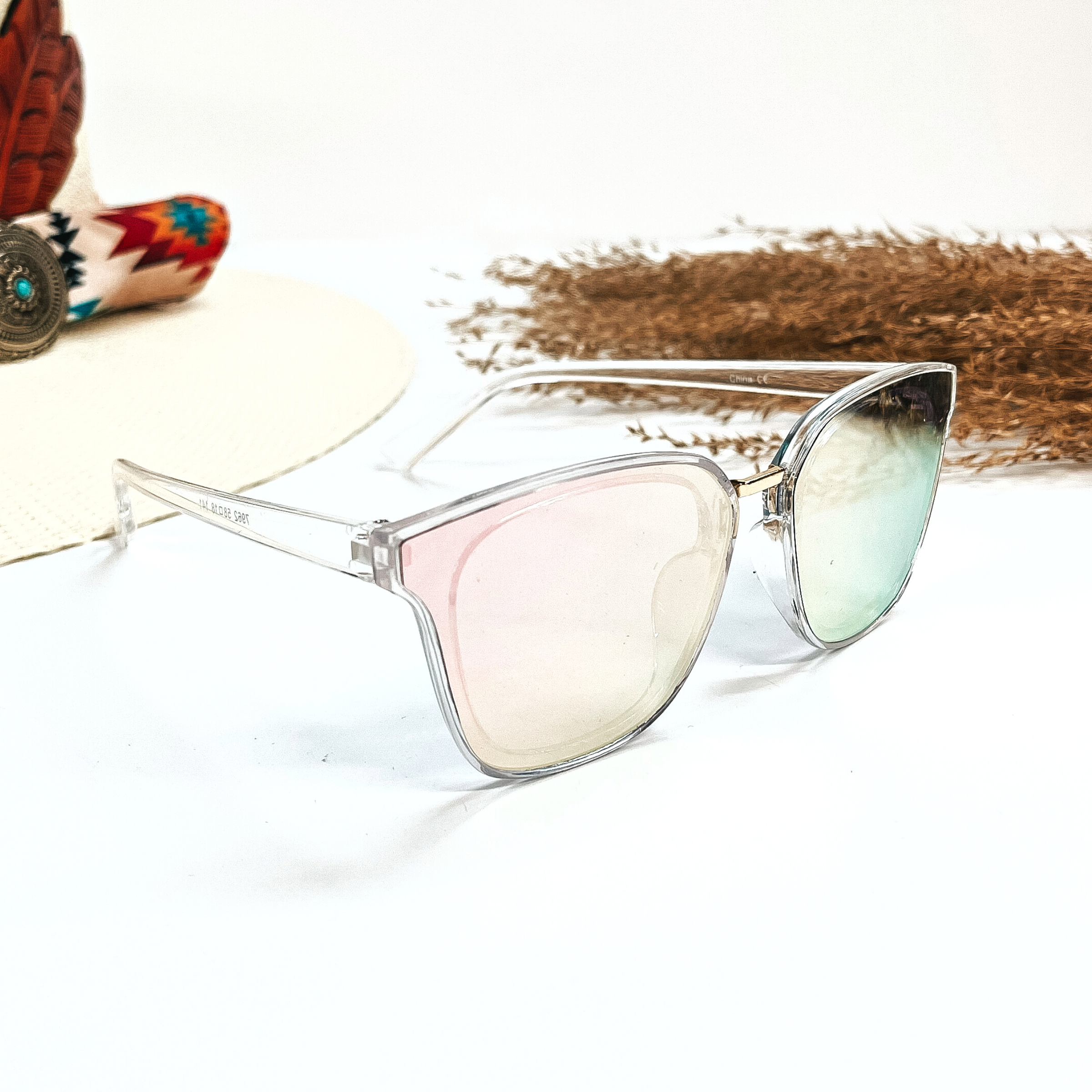 This is a pair of wayfarer style sunglasses with a transparent/clear frame,  coral/light green lense. These sunglasses are taken on a white  background, there is a straw hat with a colorful aztec print hat band and a brown plant  in the side as decor.