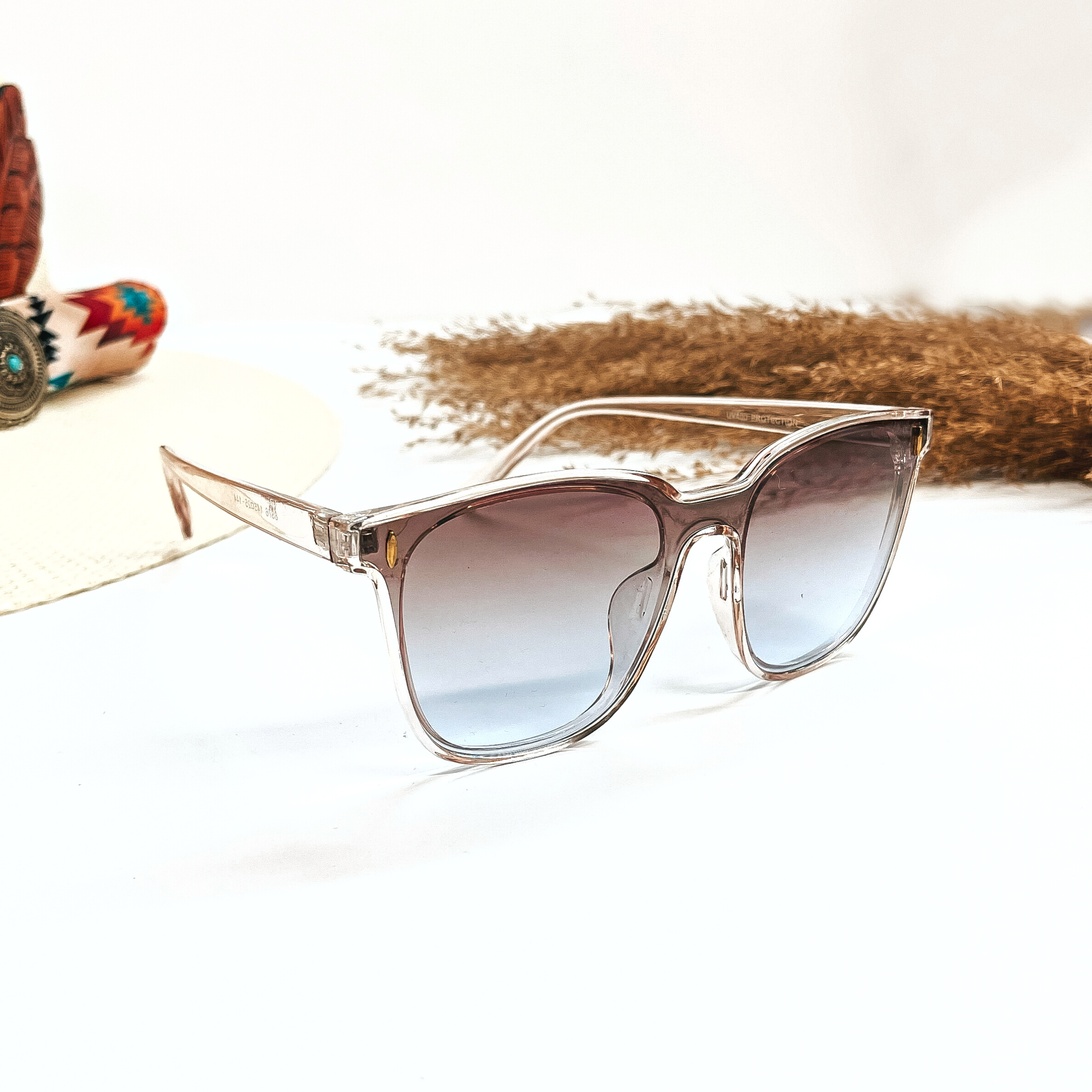 This is a pair of wayfarer style sunglasses with a transparent/clear light pink frame,  brown/light grey gradient lense with a gold tone detailing. These sunglasses are taken on a white  background, there is a straw hat with a colorful aztec print hat band and a brown plant  in the side as decor.