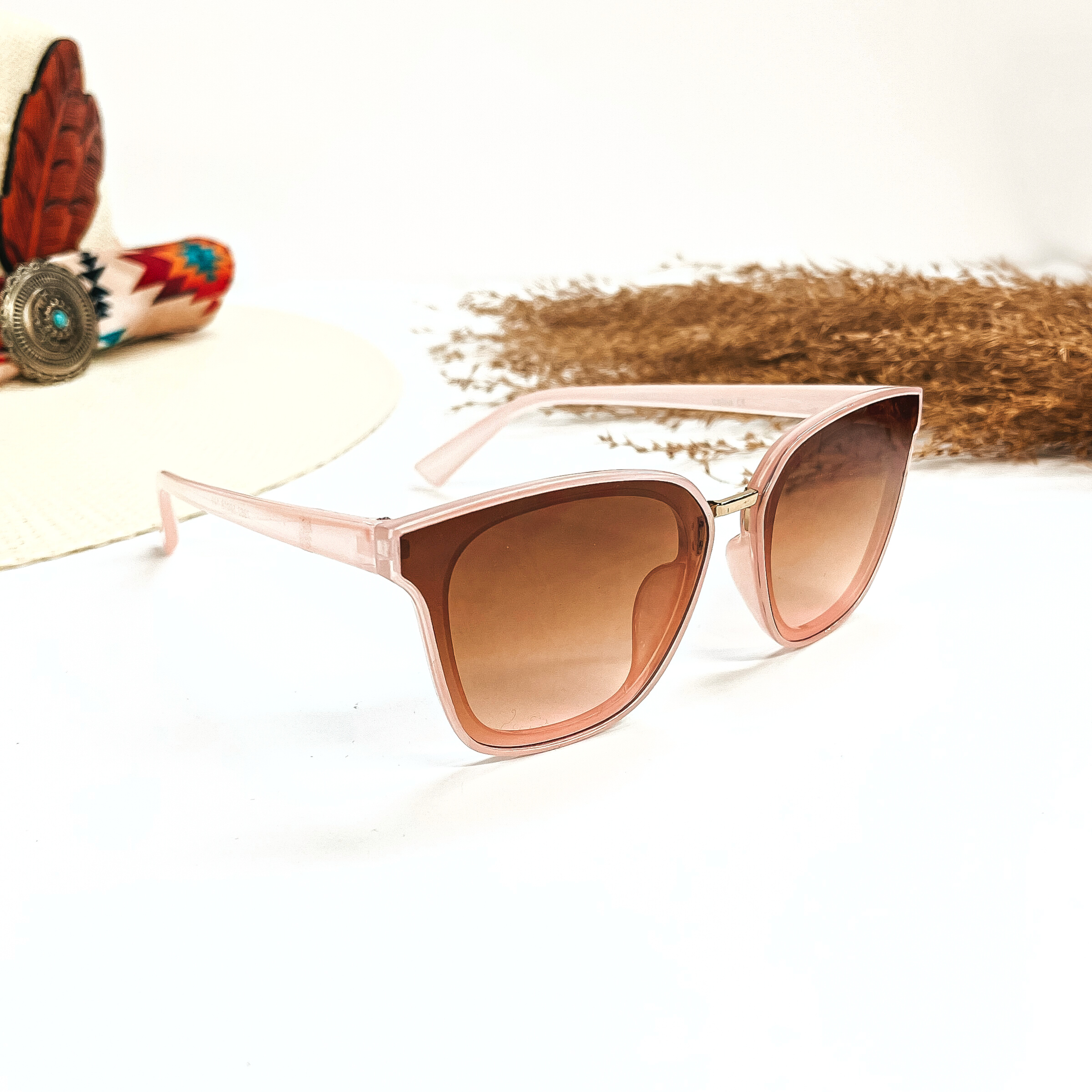 This is a pair of wayfarer style sunglasses with a transparent pink frame, brown/light  pink gradient lense with a gold tone connector. These sunglasses are taken on a white  background, there is a straw hat with a colorful aztec print hat band and a brown plant  in the side as decor.