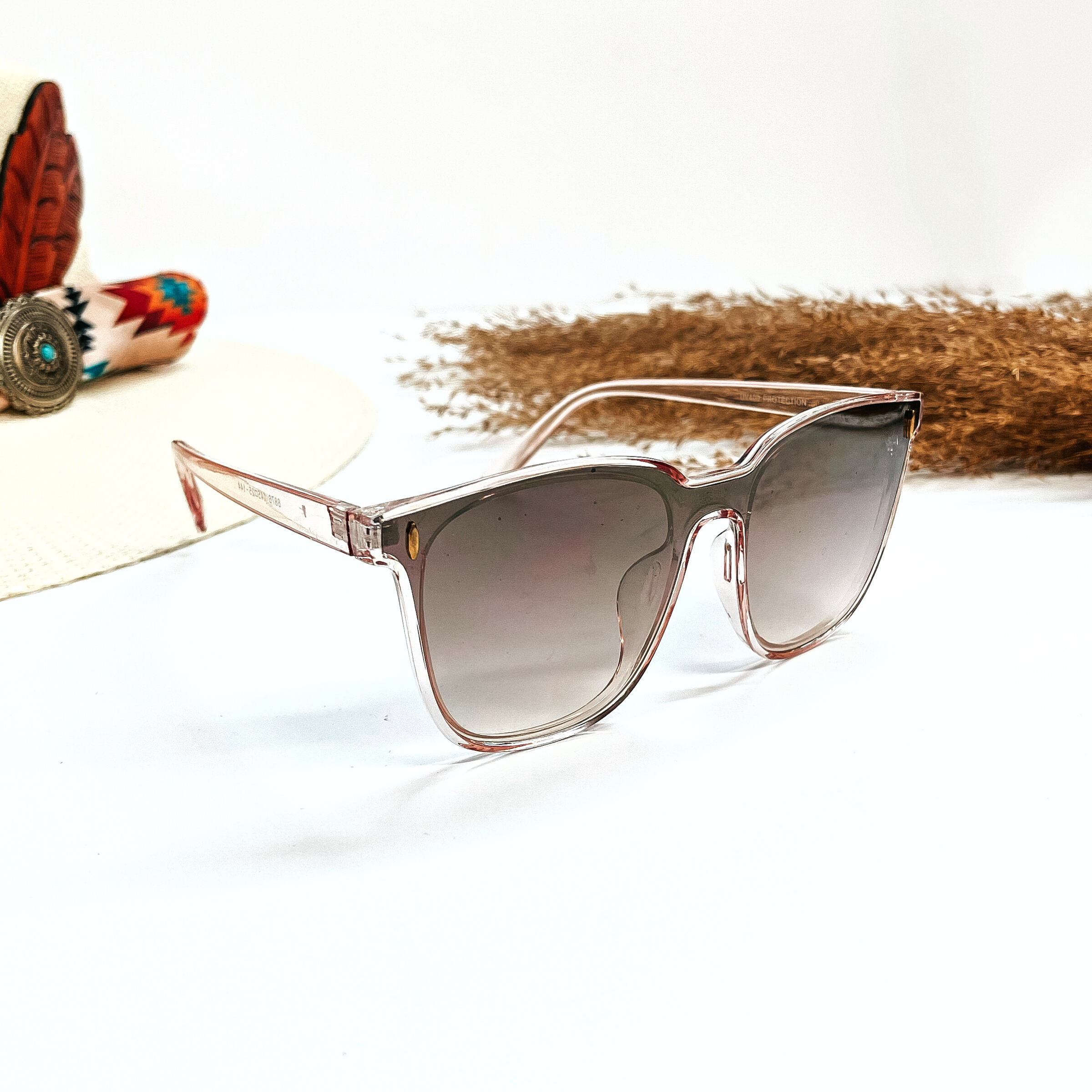 This is a pair of wayfarer style sunglasses with a transparent/clear light pink frame,  grey/light pink gradient lense with a gold tone detailing. These sunglasses are taken on a white  background, there is a straw hat with a colorful aztec print hat band and a brown plant  in the side as decor.