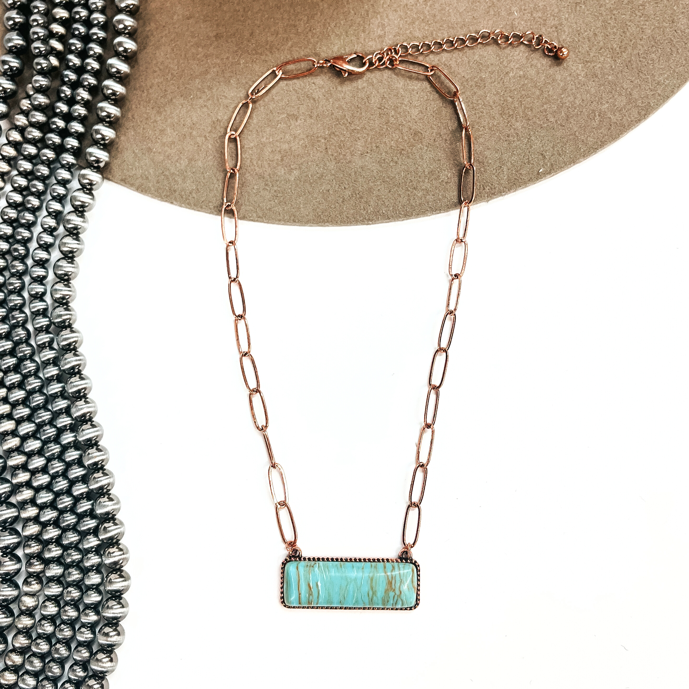 This is a copper tone thin link chain with a rectangle bar pendant in the center. The  rectangle bar pendant has an agate stone in turquoise. This necklace is laying on a white  background and light brown felt hat brim, with silver Navajo beads in the side as decor.
