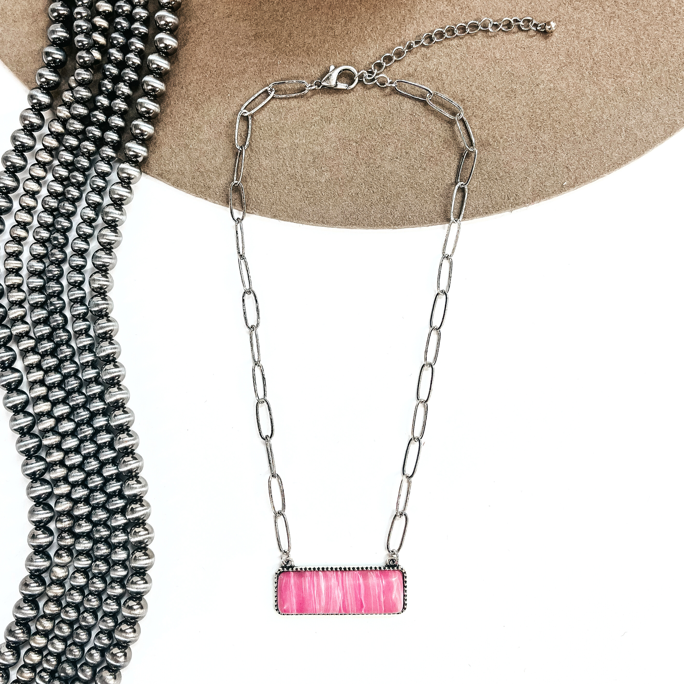 This is a silver tone thin link chain with a rectangle bar pendant in the center. The  rectangle bar pendant has an agate stone in pink. This necklace is laying on a white  background and light brown felt hat brim, with silver Navajo beads in the side as decor.