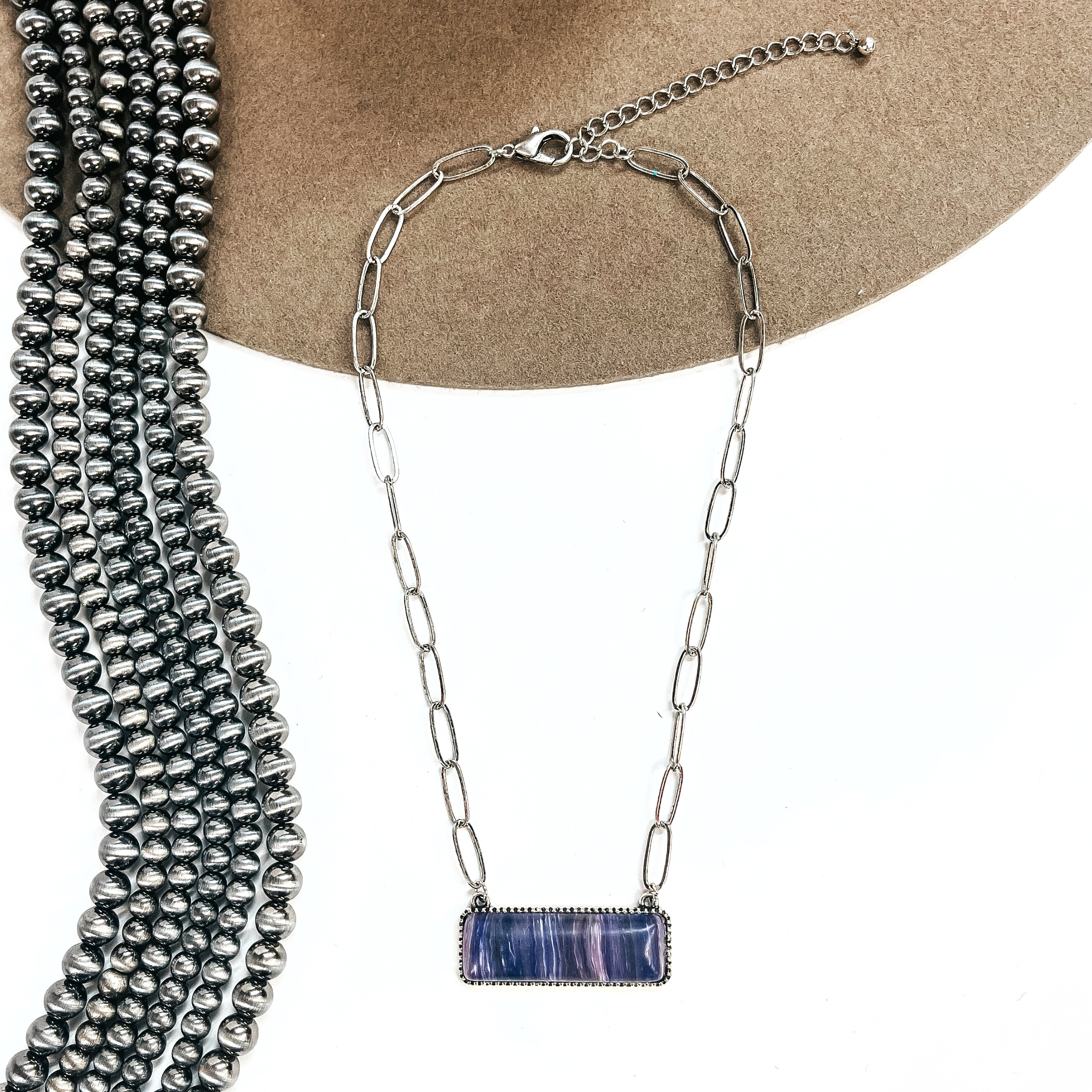 This is a silver tone thin link chain with a rectangle bar pendant in the center. The  rectangle bar pendant has an agate stone in purple. This necklace is laying on a white  background and light brown felt hat brim, with silver Navajo beads in the side as decor.
