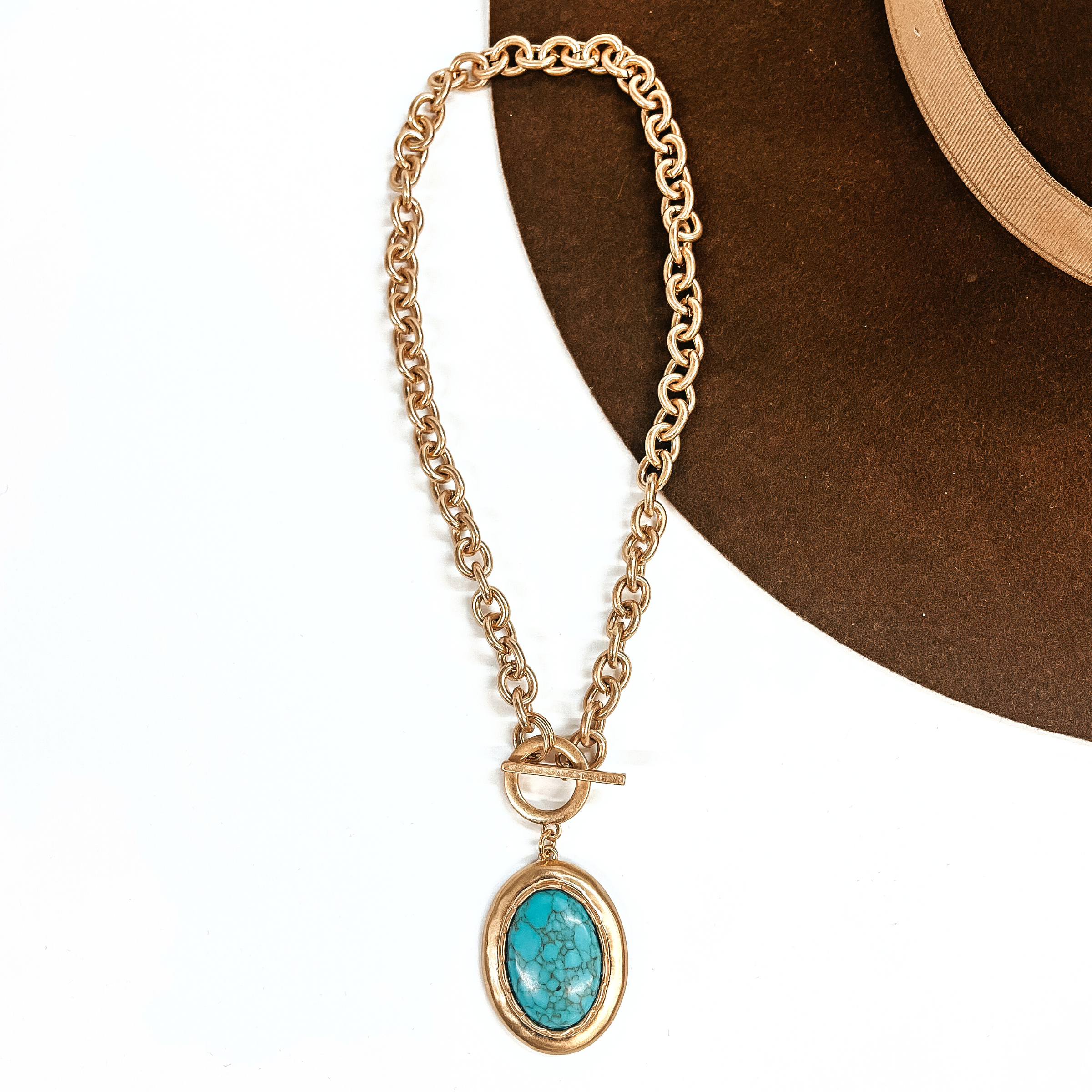 This is a matte gold tone, thick link chain necklace with a front toggle clasp. There is  an oval pendant drop, a turquoise stone in a gold setting. This necklace is taken on a white background and on a dark brown felt hat brim.
