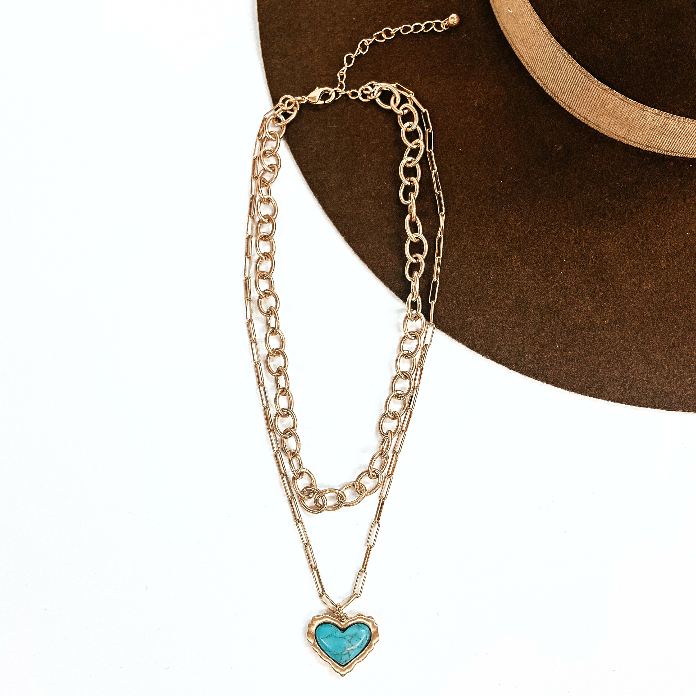 This is a two strand matte gold tone chained necklace with a heart pendant. The small  strand is a small,thick link chain and the long strand is a thin small linked chain with  a turquoise heart stone in a gold setting. This necklace is taken on a white background  and on a dark brown felt hat brim.