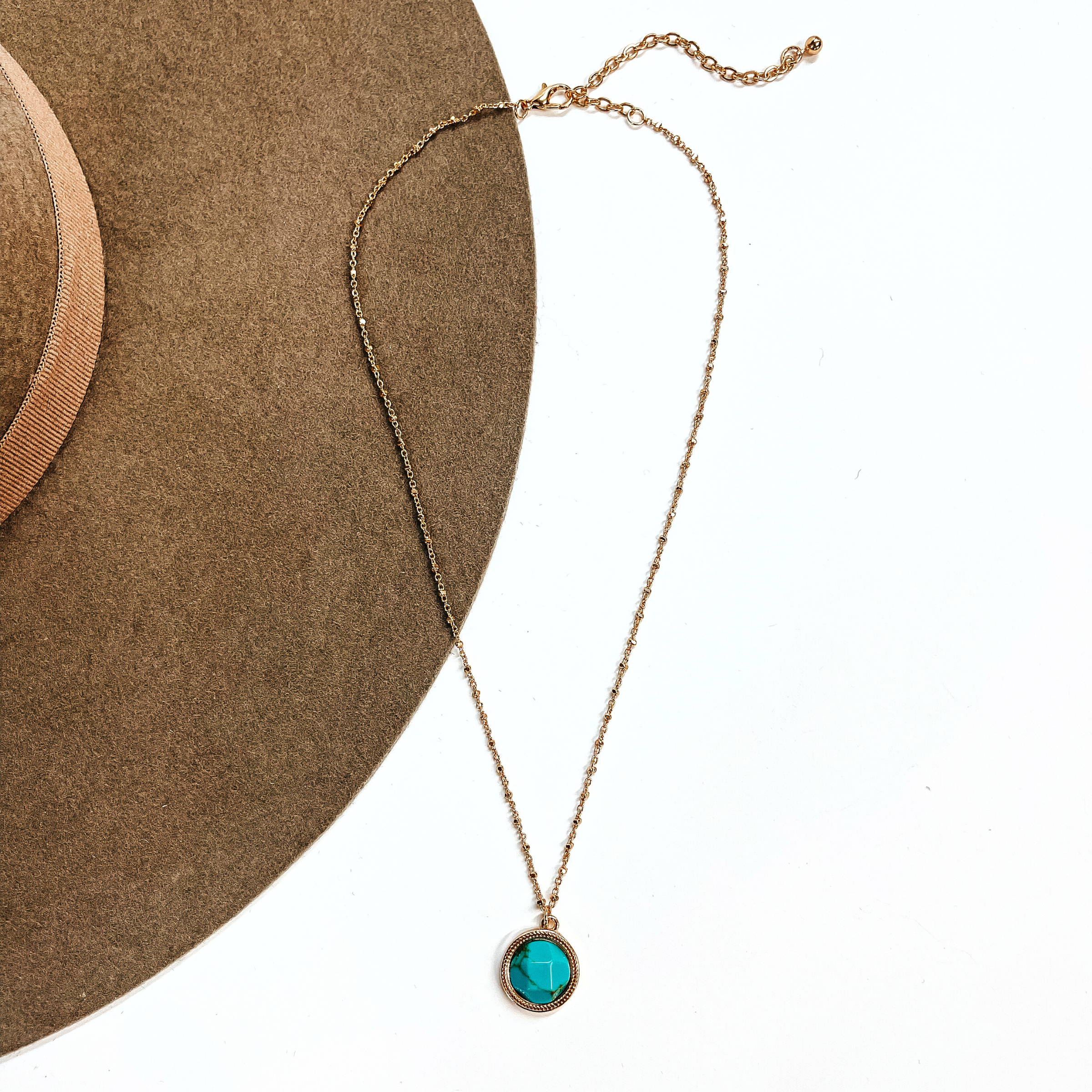 This is a thin gold tone chain necklace with a small circle pendant in the center. The  pendant has a turqouise stone shaped as a circle, the stone is in a gold setting. This  necklace is laying on a white background and on a light brown felt hat brim.