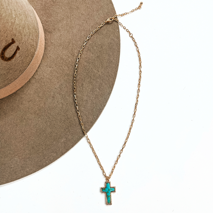 This is a thin gold tone chain necklace with a small cross pendant in the center. The  pendant has a turqouise stone shaped as a cross, the stone is in a gold setting. This  necklace is laying on a white background and on a light brown felt hat brim.