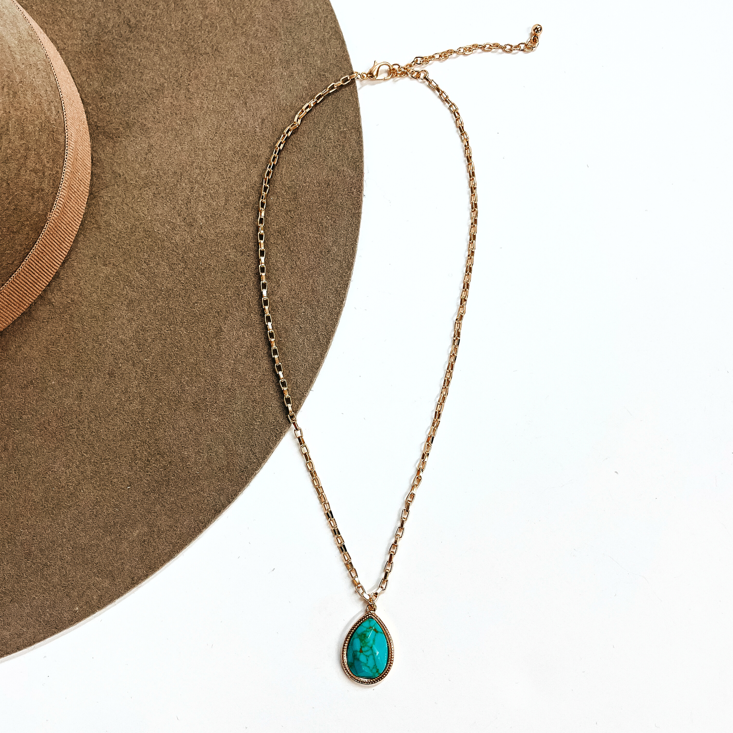 This is a thin gold tone chain necklace with a small teardrop pendant in the center. The  pendant has a turqouise stone shaped as a teardrop, the stone is in a gold setting. This  necklace is laying on a white background and on a light brown felt hat brim.