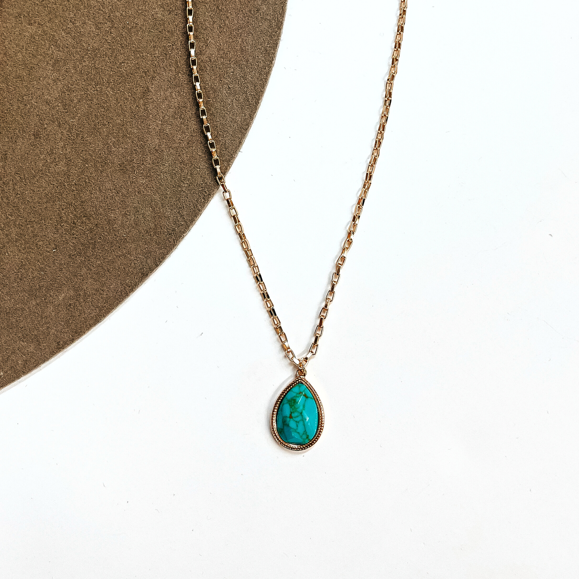 This is a thin gold tone chain necklace with a small teardrop pendant in the center. The  pendant has a turqouise stone shaped as a teardrop, the stone is in a gold setting. This  necklace is laying on a white background and on a light brown felt hat brim.