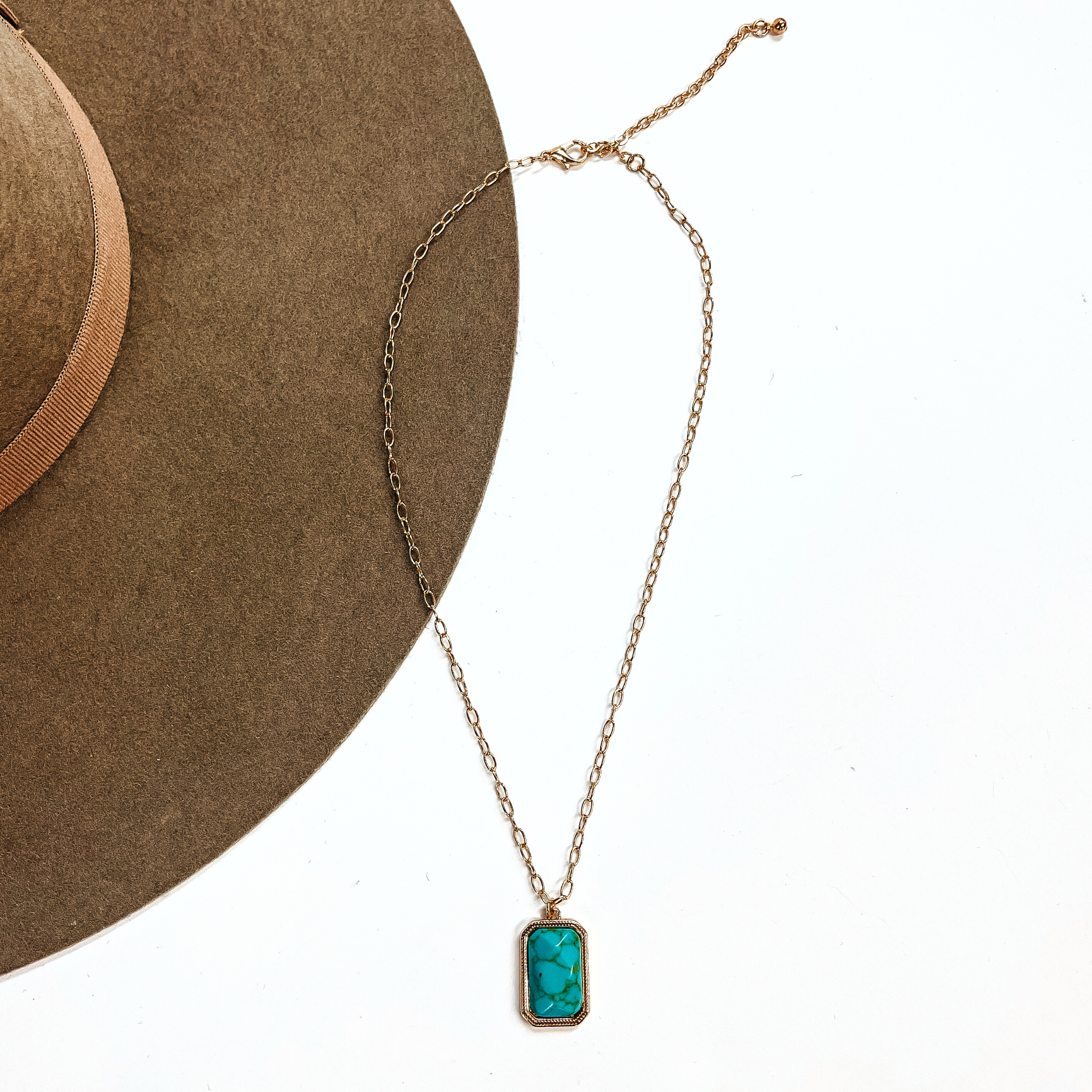 This is a thin gold tone chain necklace with a rectangle pendant in the center. The  pendant has a turqouise stone shaped as a rectangle, the stone is in a gold setting. This  necklace is laying on a white background and on a light brown felt hat brim.