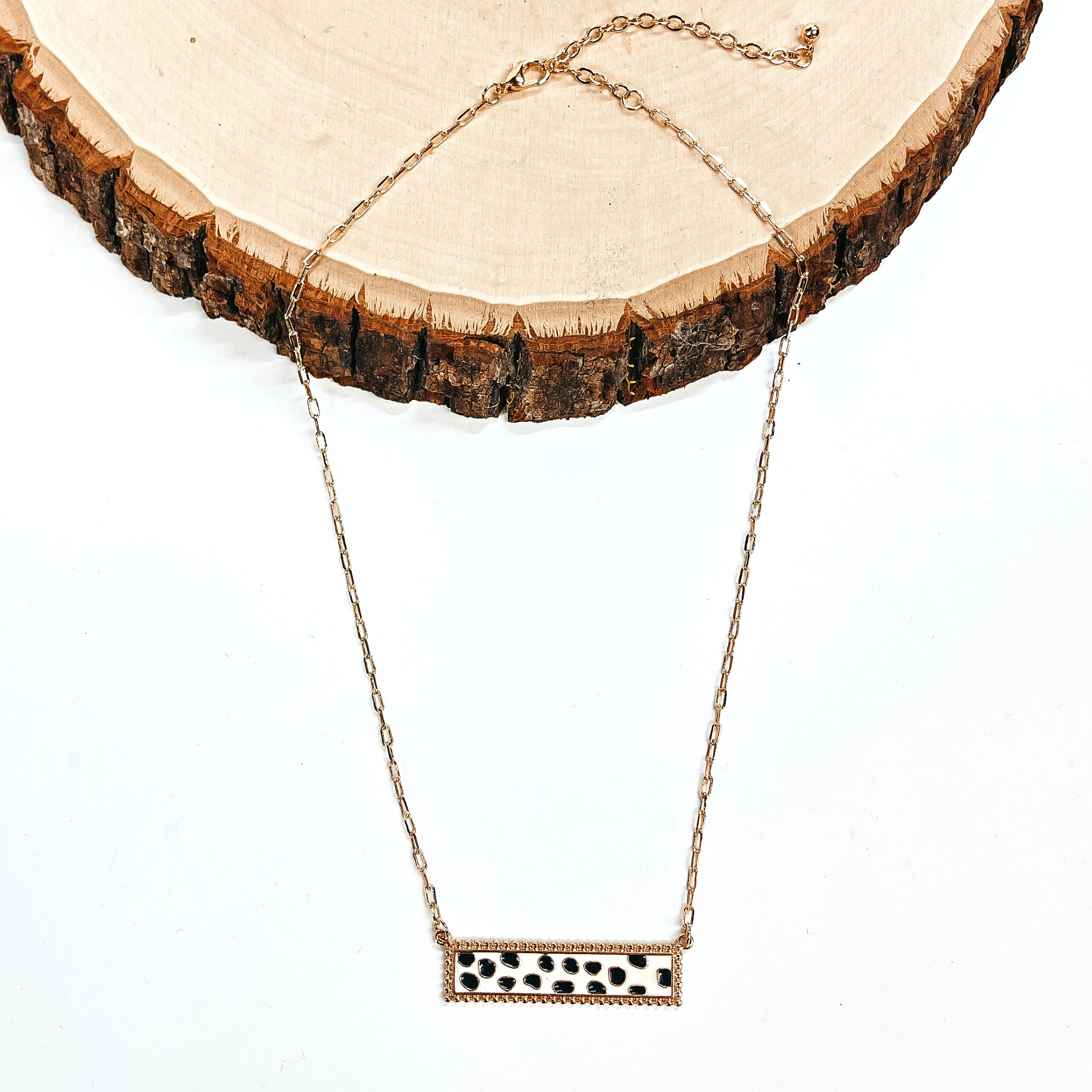 This is a thin gold tone chain necklace with a rectangle bar pendant in black and white  dotted print. The black and white dotted print rectangle bar is in a gold tone setting. This necklace is taken laying on a white background and a slab of wood.
