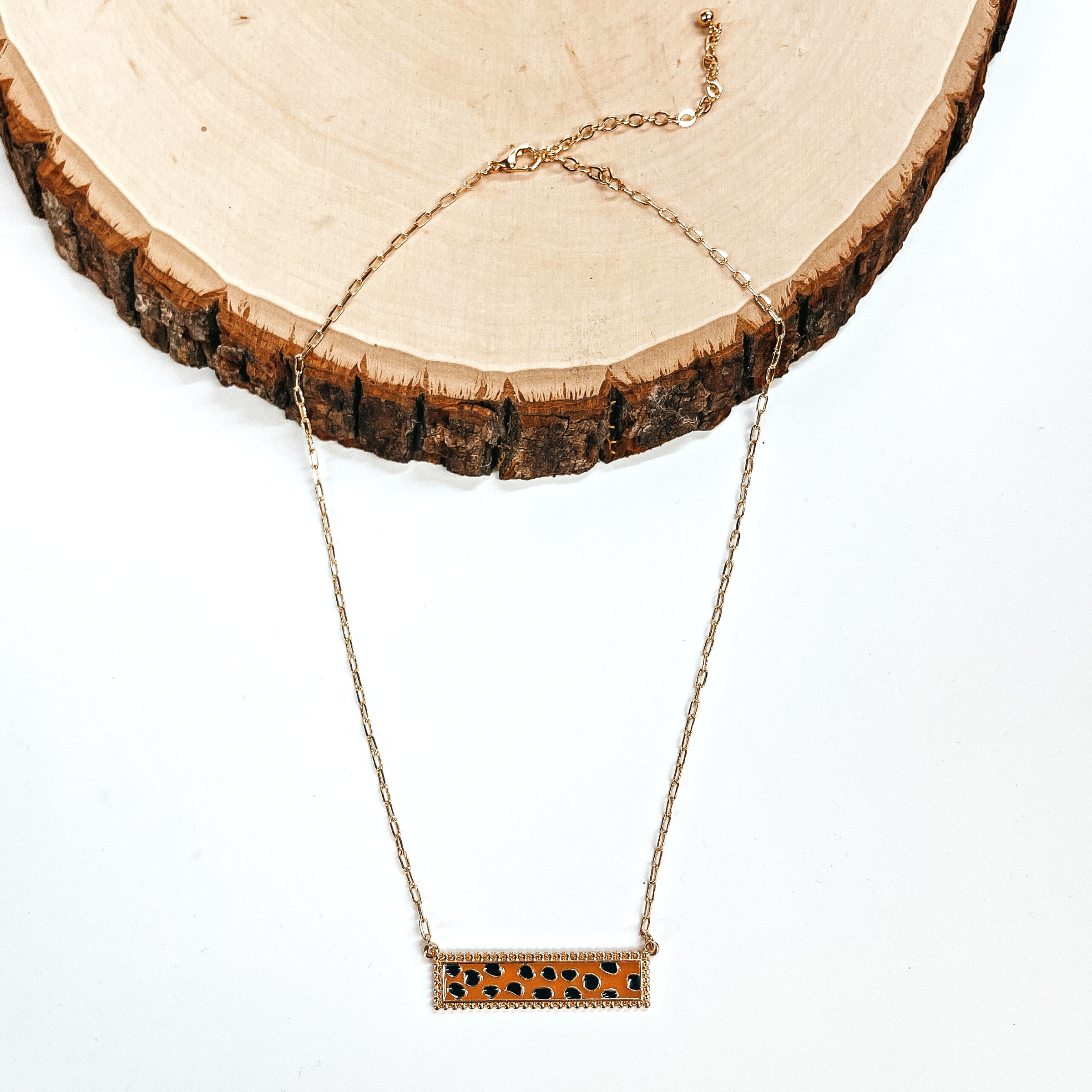 This is a thin gold tone chain necklace with a rectangle bar pendant in black and brown  dotted print. The black and brown dotted print rectangle bar is in a gold tone setting. This necklace is taken laying on a white background and a slab of wood.