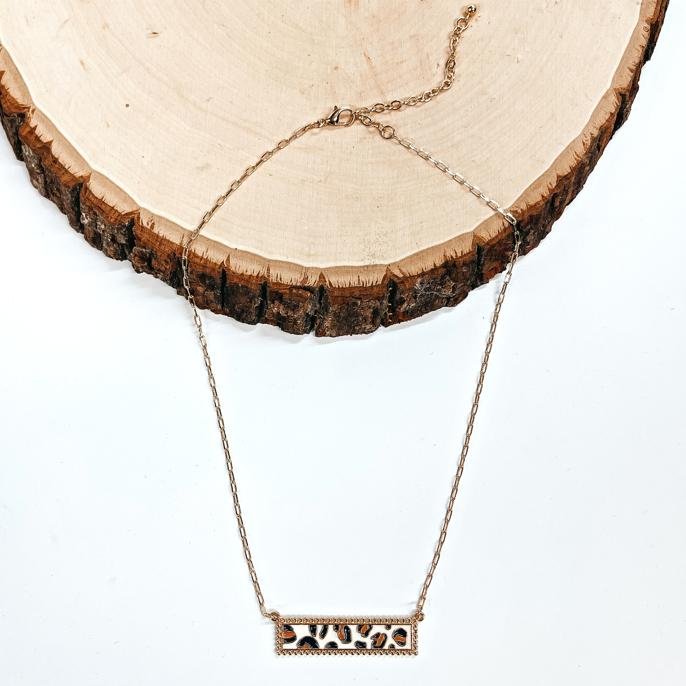 This is a thin gold tone chain necklace with a rectangle bar pendant in black,brown, and  white/ivory leopard print. The black, brown, white/ivory leopard print rectangle bar is  in a gold tone setting. This necklace is taken laying on a white background and a slab of wood.