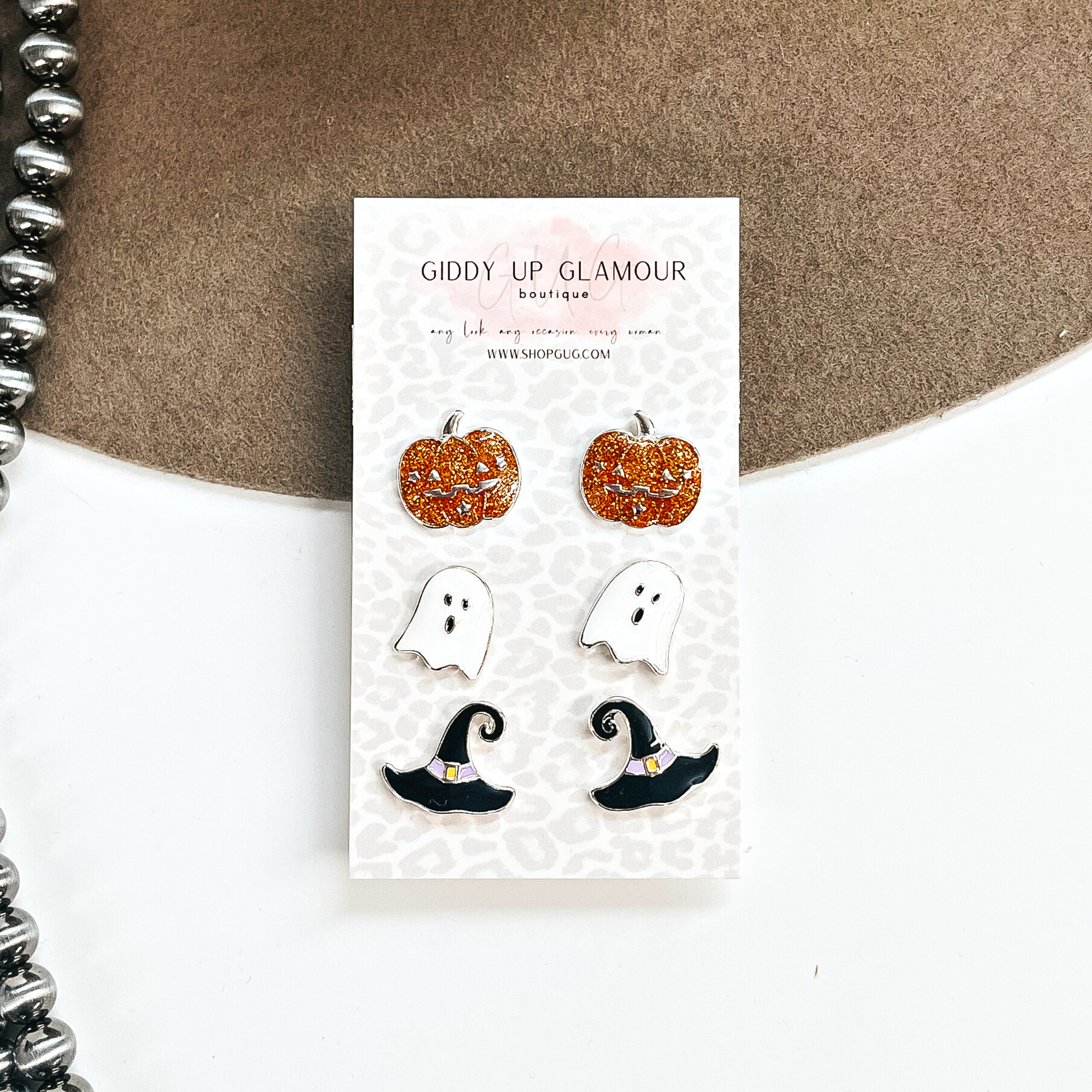 This is a set of three stud earrings with Halloween charms. From top to bottom; pumpkins  in orange glitter in a silver setting, white ghost with a black eyes and mouth in a  silver setting, and black witch hat's with a purple band in a silver setting. These  earrings are places on a Giddy Up Glamour card, they are taken on a brown hat brim  and white background with silver Navajo beads in the side as decor.