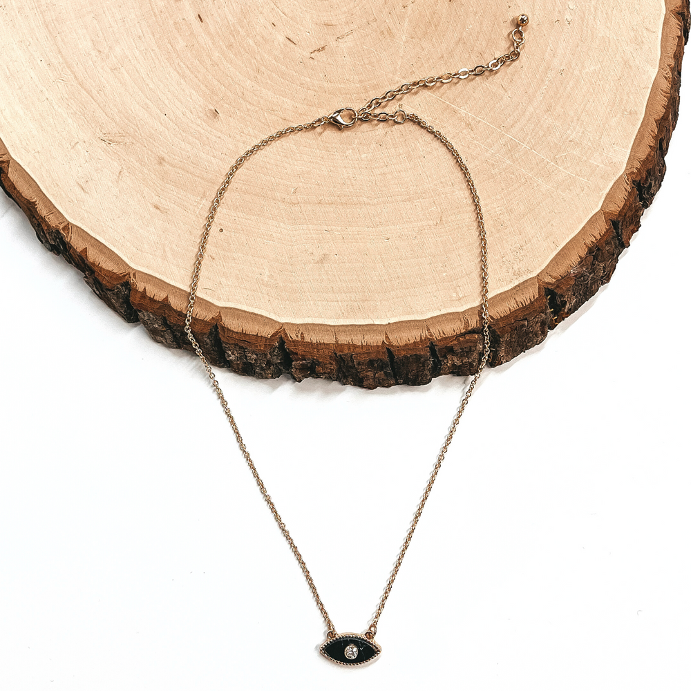 This is a small thin gold necklace with a black evil eye charm in the middle,  with a small clear crystal in the charm. This necklace is laying on a white  background and on a slab of wood.