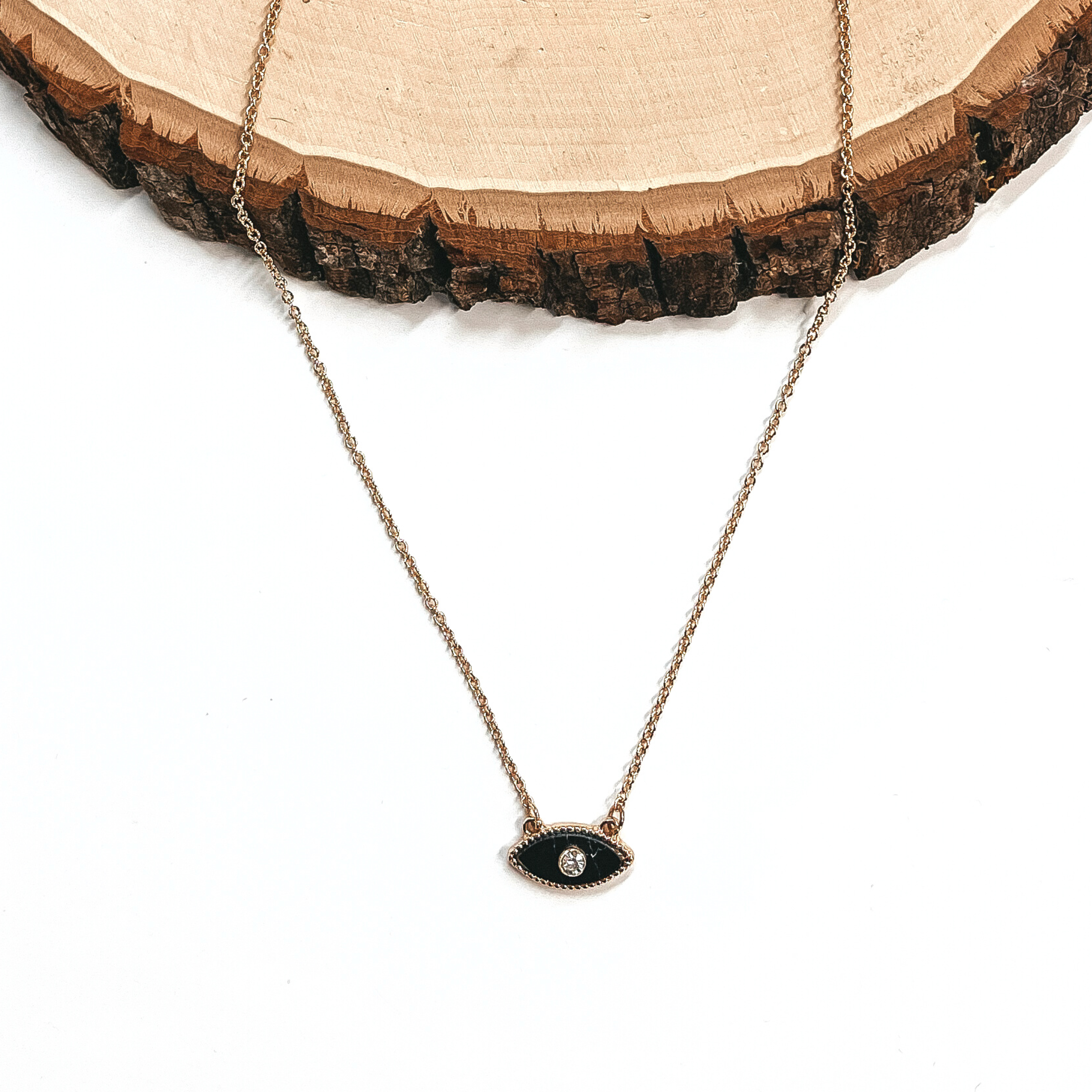 This is a small thin gold necklace with a black evil eye charm in the middle,  with a small clear crystal in the charm. This necklace is laying on a white  background and on a slab of wood.