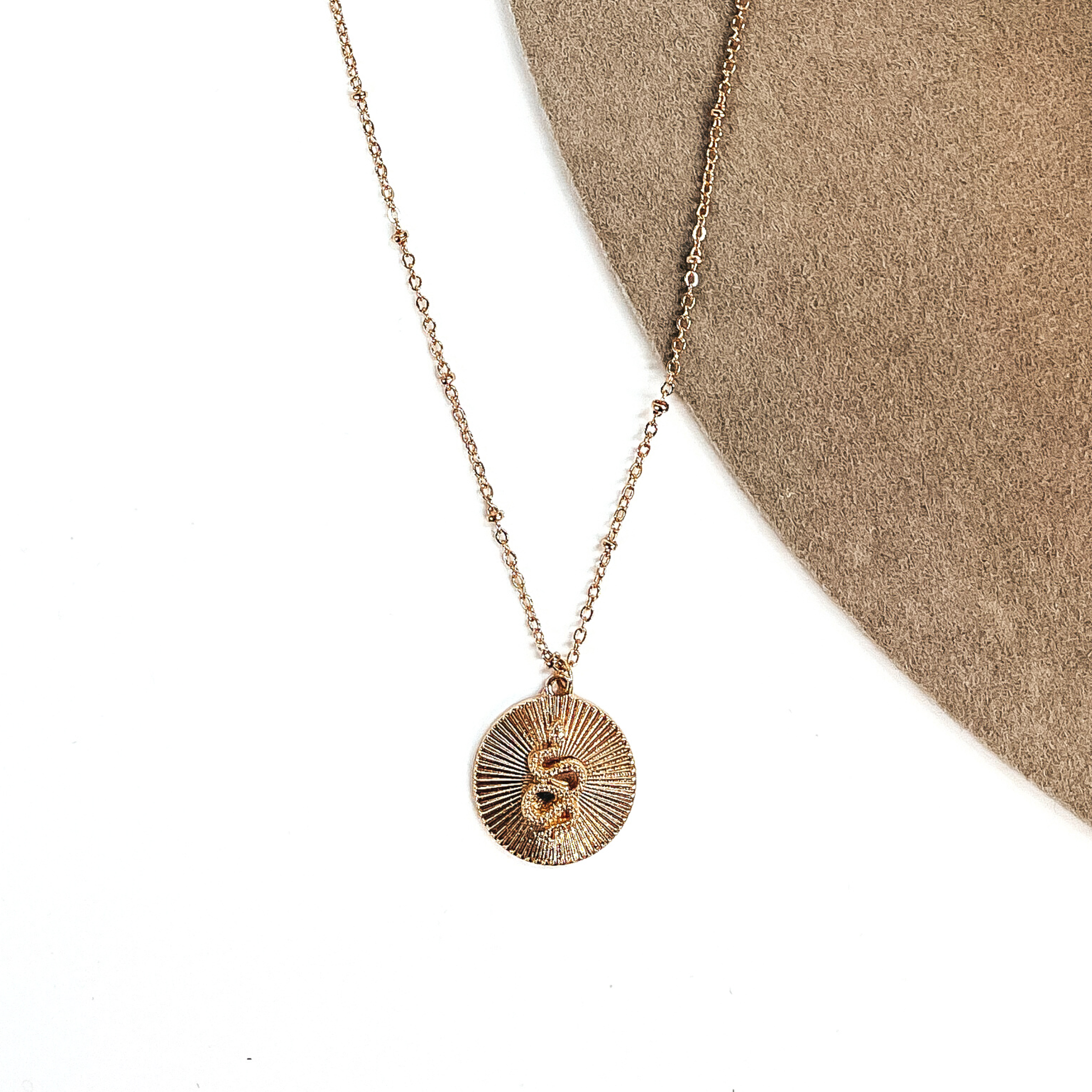 This is a small thin gold chain necklace with a small circle pendant in  textured sunburst. The pendant has a small snake in the center. This necklace  is taken on a white background and on a beige felt hat.