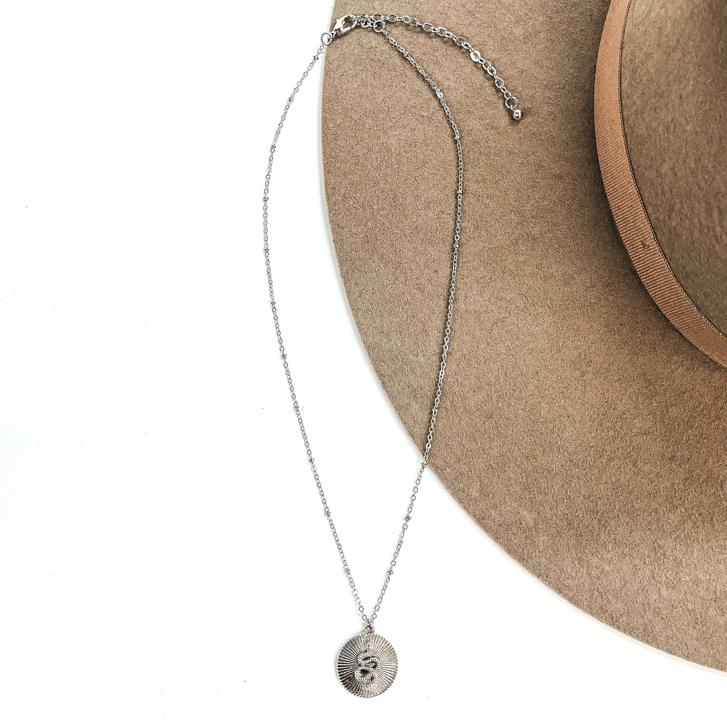 This is a small thin silver chain necklace with a small circle pendant in  textured sunburst. The pendant has a small snake in the center. This necklace  is taken on a white background and on a beige felt hat.