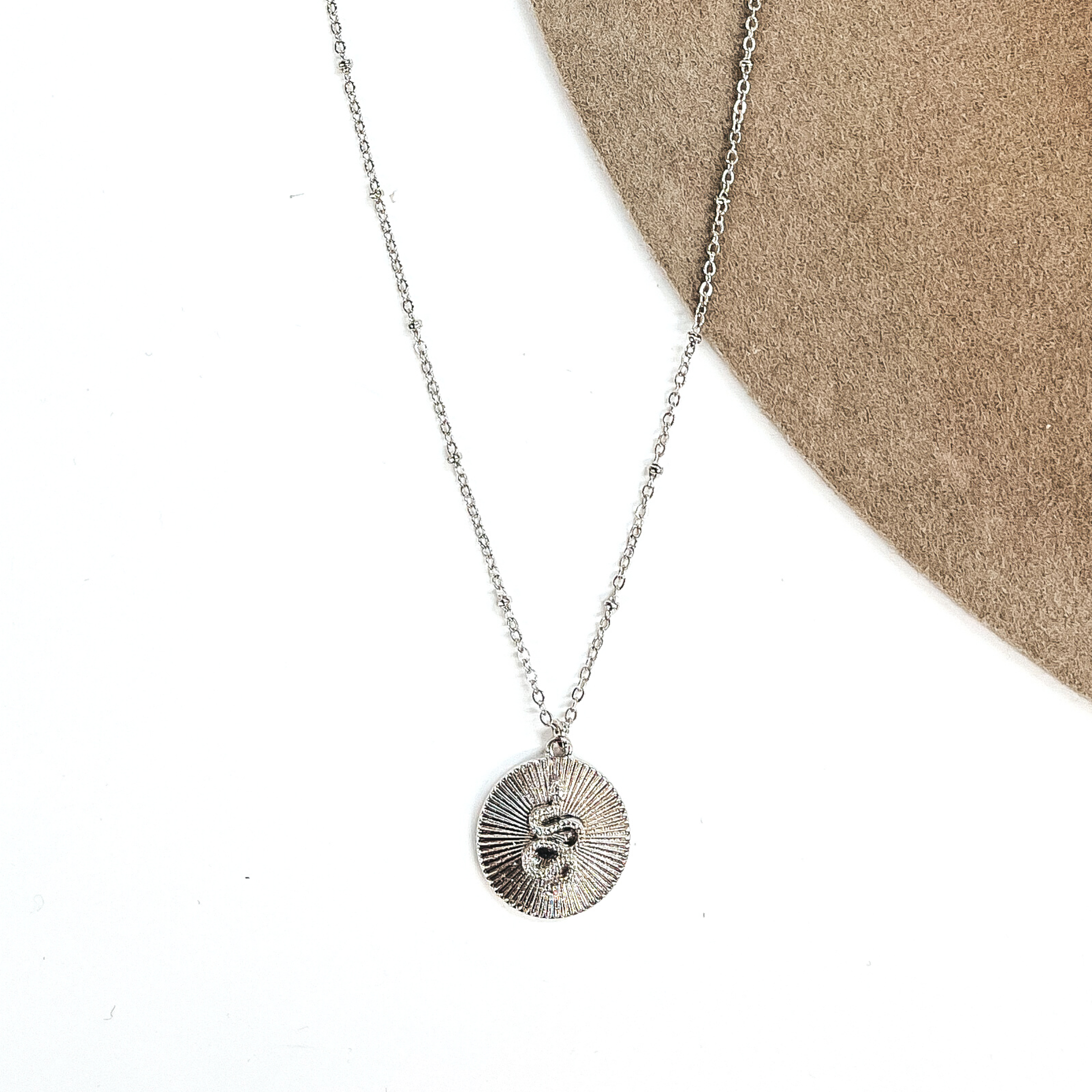 This is a small thin silver chain necklace with a small circle pendant in  textured sunburst. The pendant has a small snake in the center. This necklace  is taken on a white background and on a beige felt hat.