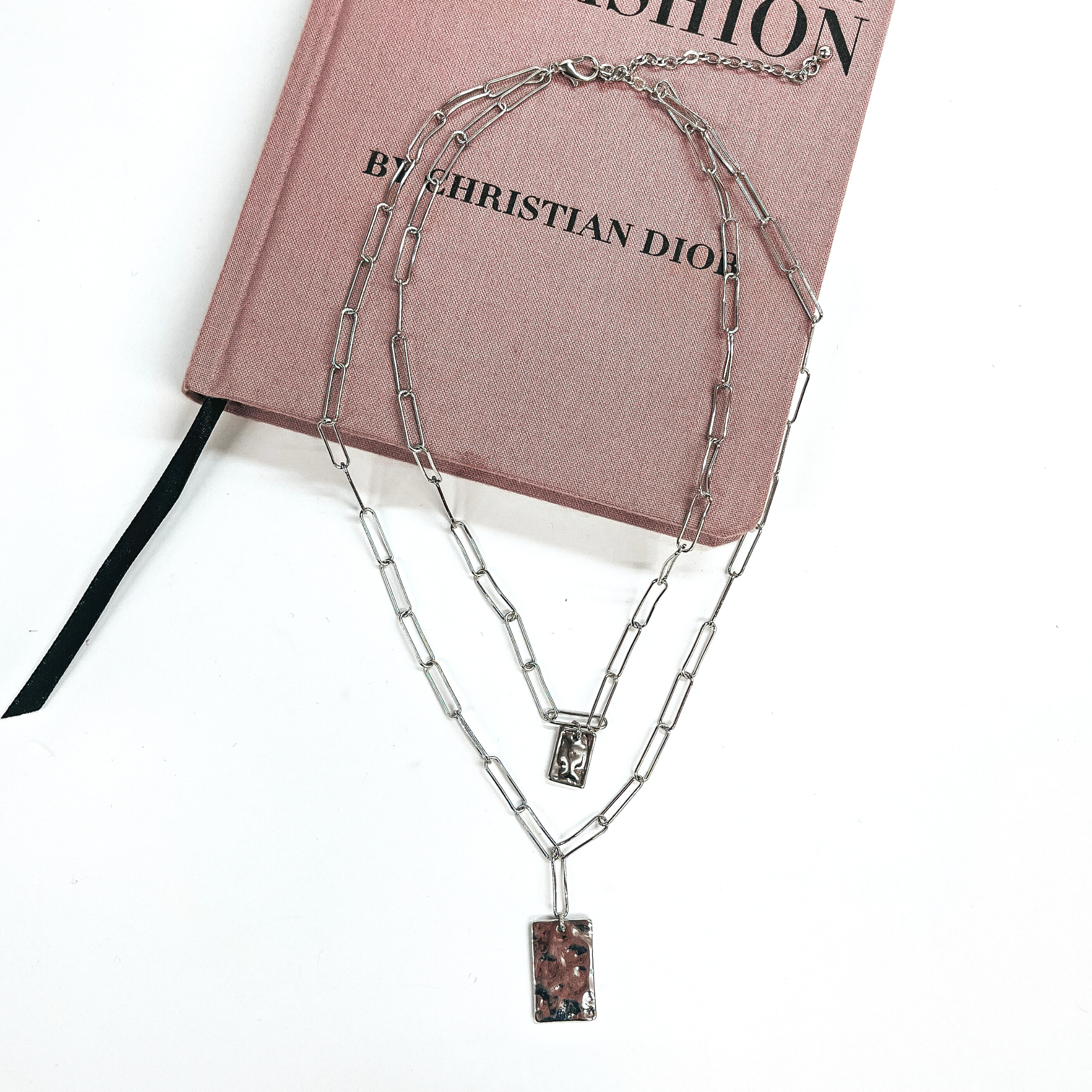 This is a two strand thin paperclips necklace in silver with two textured  rectangle pendants in each strand. The longest strand has a bigger rectangle  pendant and the smaller strand has a smaller pendant. This necklace is taken  on a purple/pink fashion book and on a white background.