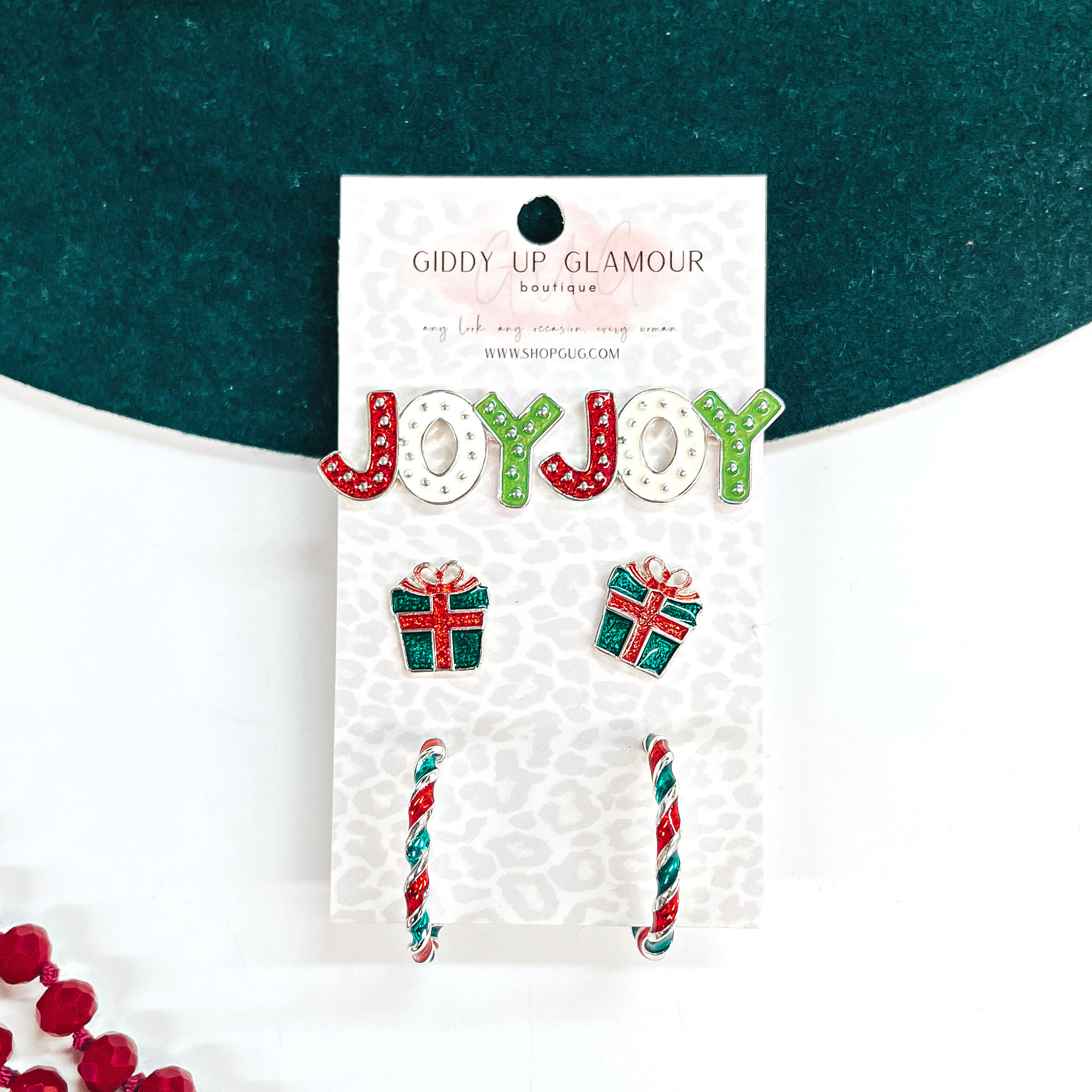 This is a set of three earrings in green, red, and white. From top to bottom;  Joy studs in red, white, and light green with small silver dots all over. The  middle pair are christmas presents in red and dark green. The bottom  earrings are small/medium twised hoops in red, gree, and silver. These  earrings are placed on a Giddy Up Glamour card, they are laying on a dark  green felt hat brim and white background with red beads in the corner as decor.